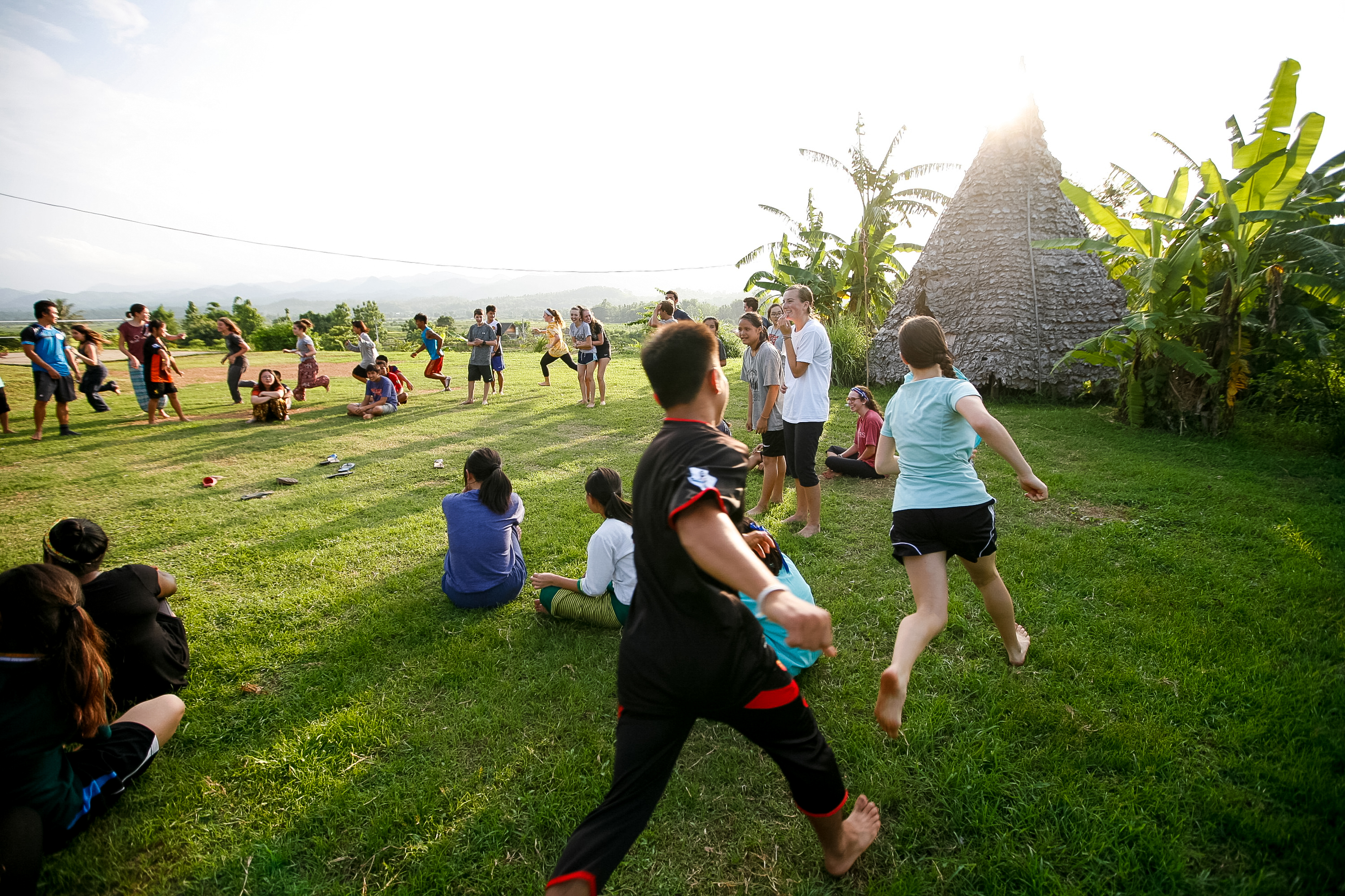 Students play a game on a grassy field near a thatched-roof structure at sunset
