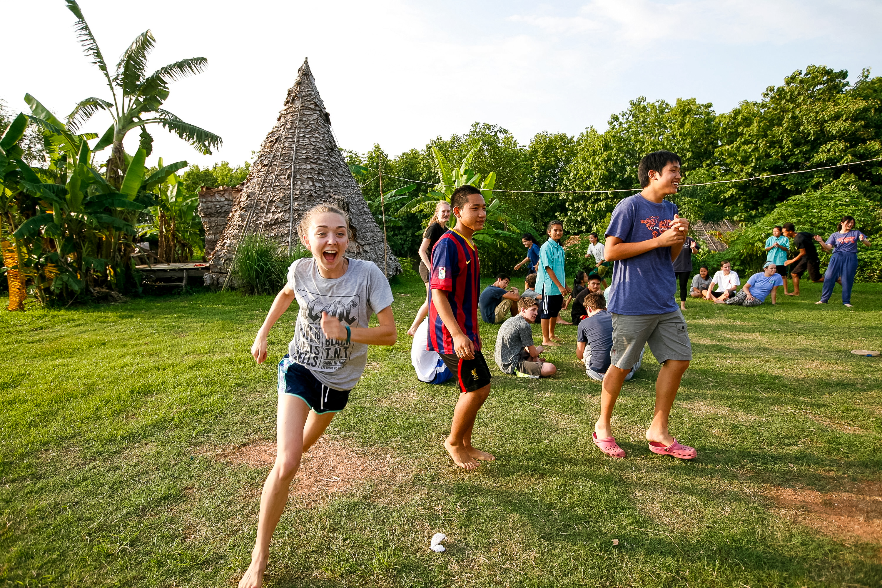 Students run and play a game on a grassy field near a thatched hut