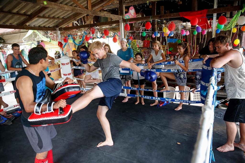 Teen practicing Muay Thai kickboxing in a vibrant training ring in Thailand, with peers watching.