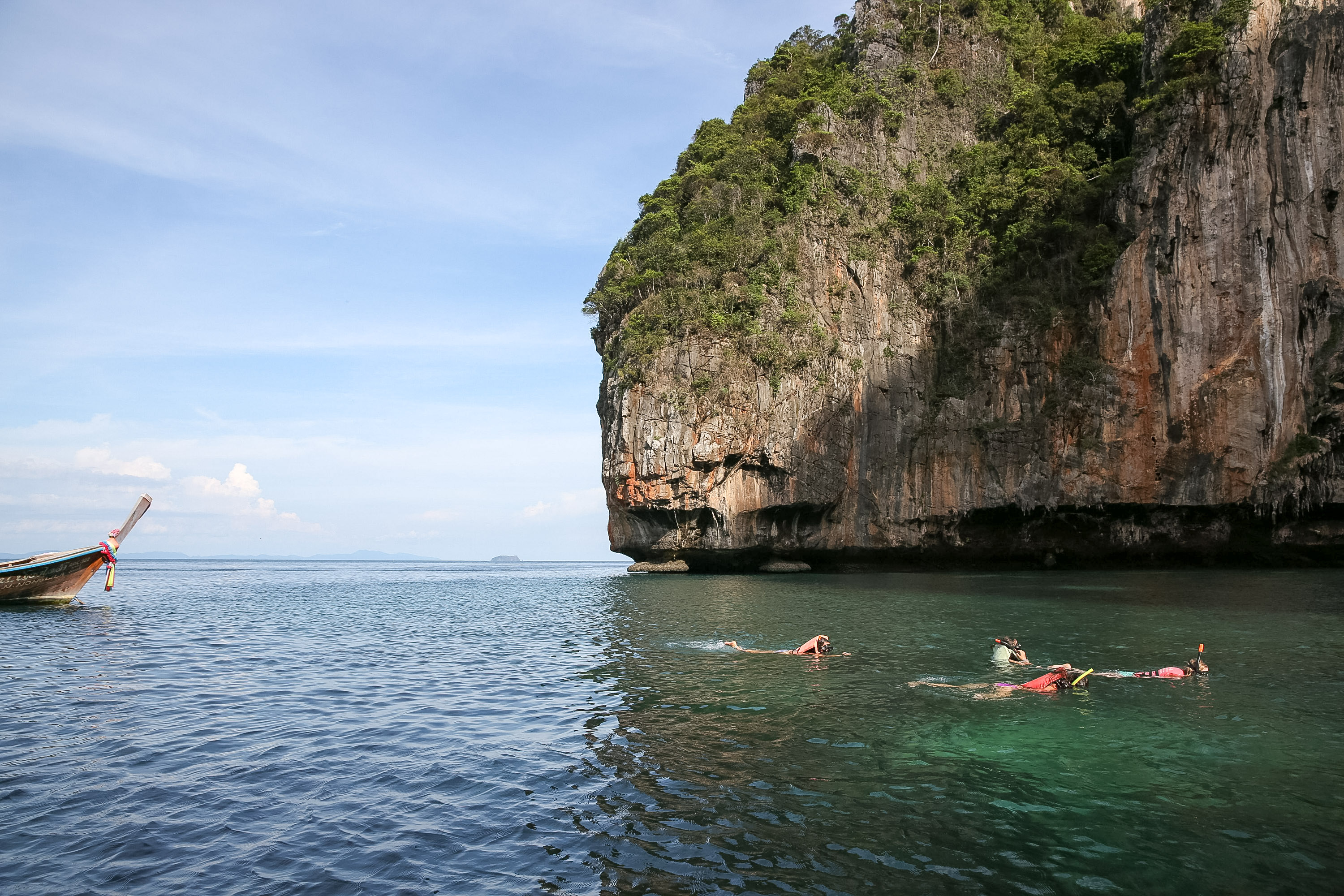 Snorkelers swim near a limestone cliff with a longtail boat nearby