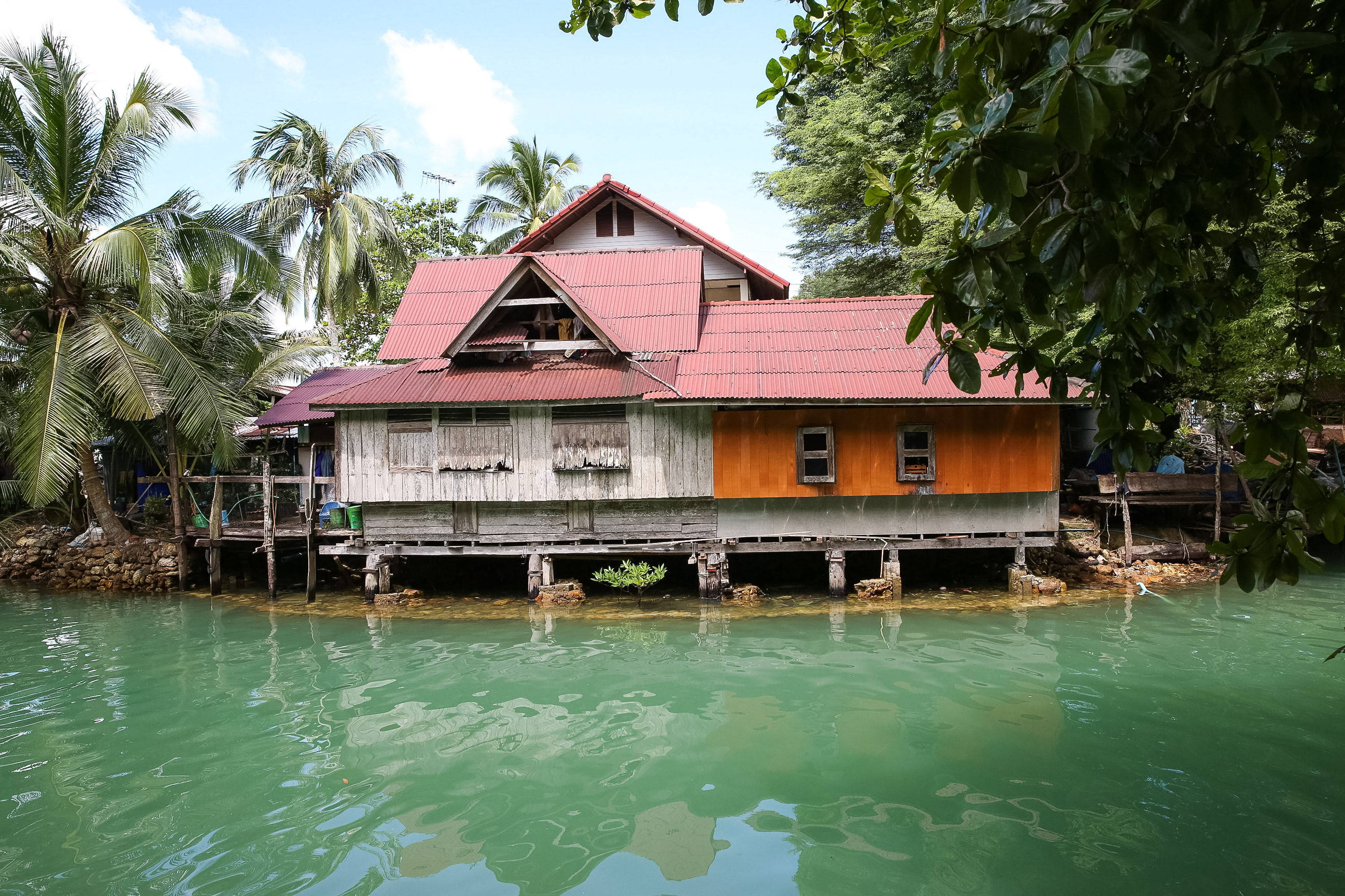 A wooden stilt house with a red roof sits over green water, framed by palm trees