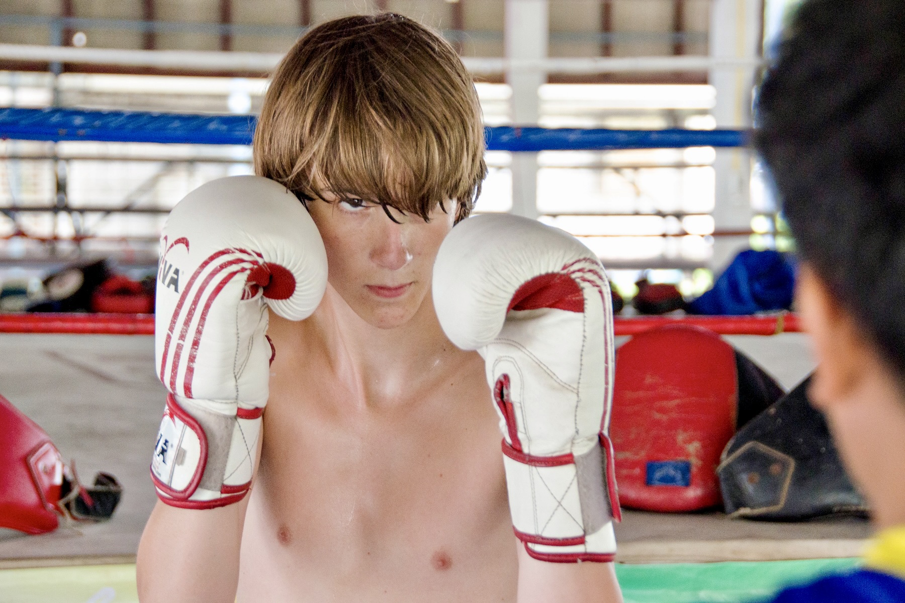 A student wearing white boxing gloves and no shirt stands in a ready stance during a Muay Thai session inside a traditional Thai boxing ring.