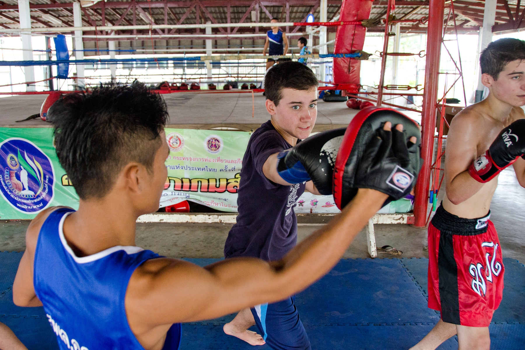 A student practices Muay Thai strikes on pads held by a trainer in a boxing ring