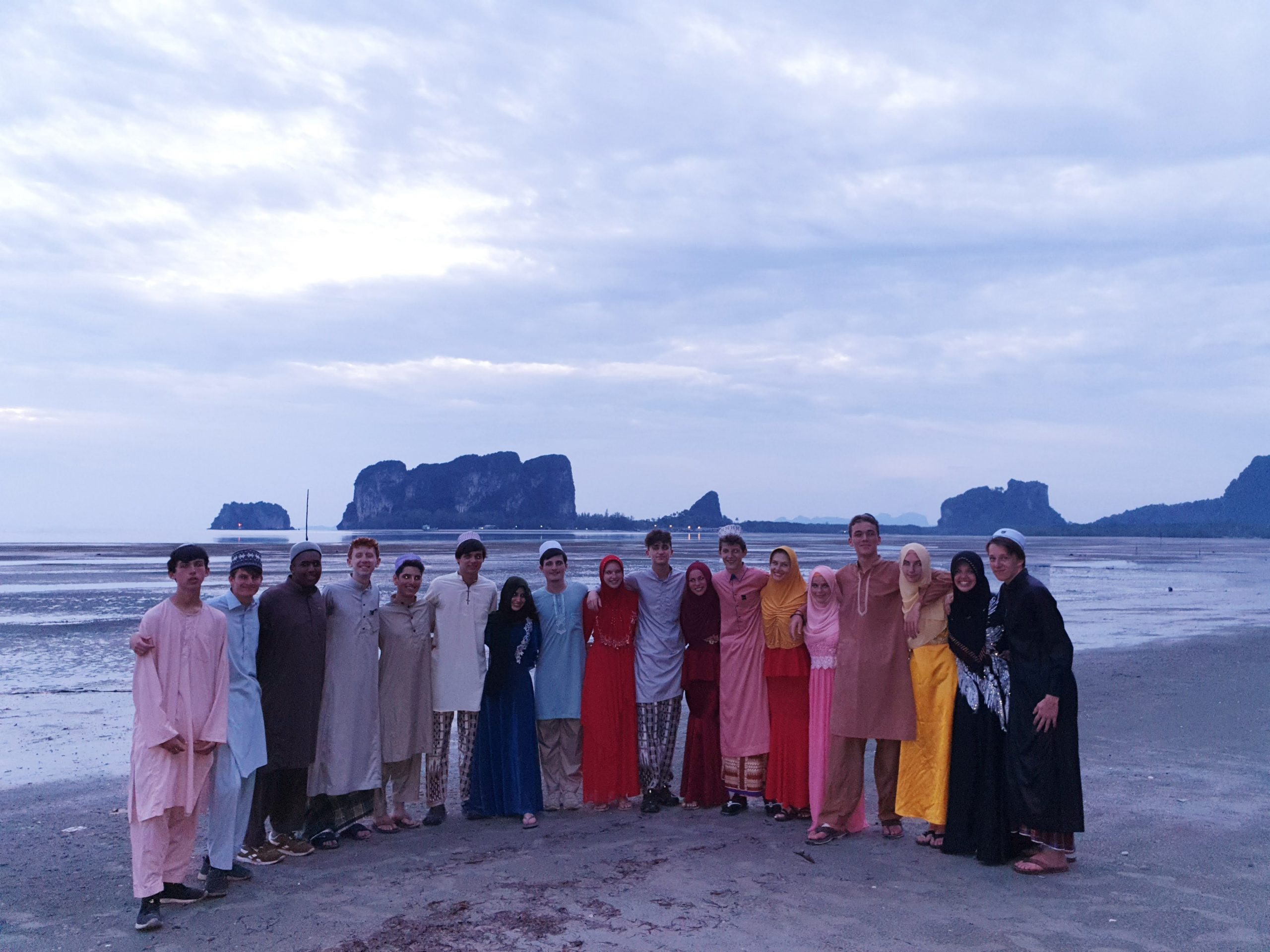 Group of students posing at the beach in Thailand with view of the ocean and rock structures in the background.