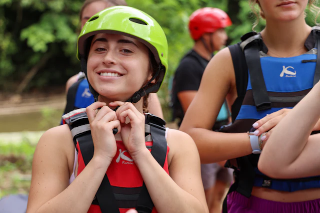 A smiling teen traveler in a green helmet fastens the chin strap of a red life jacket on a river bank, other rafters behind.