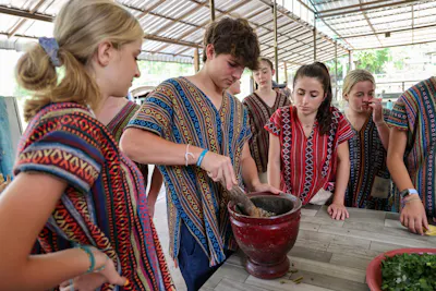 Students in traditional Thai dress grinding ingredients in a stone mortar alongside local hosts