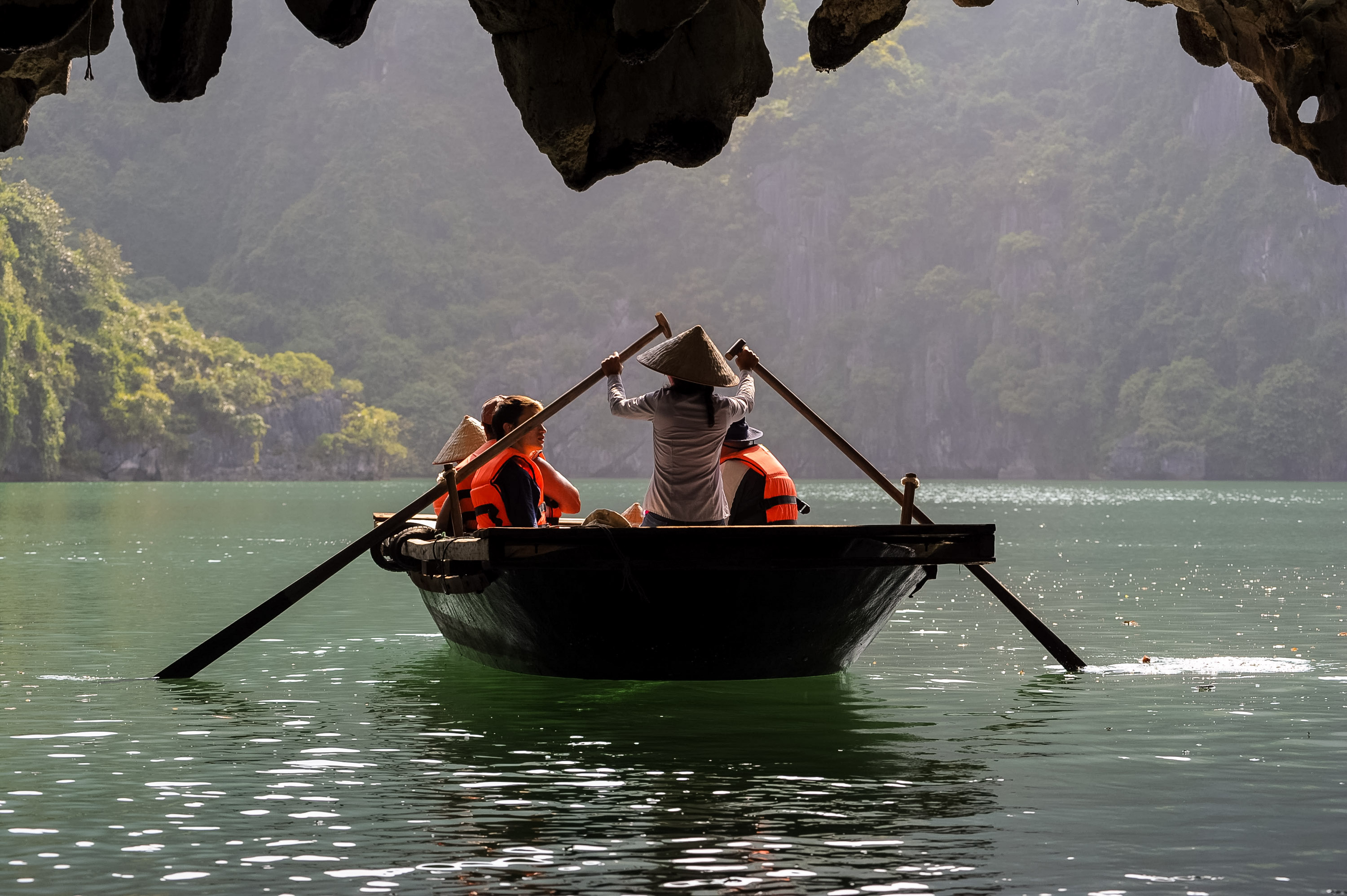 On a school trip, a rower guides a small wooden boat through limestone karst scenery as passengers in life jackets sit ahead