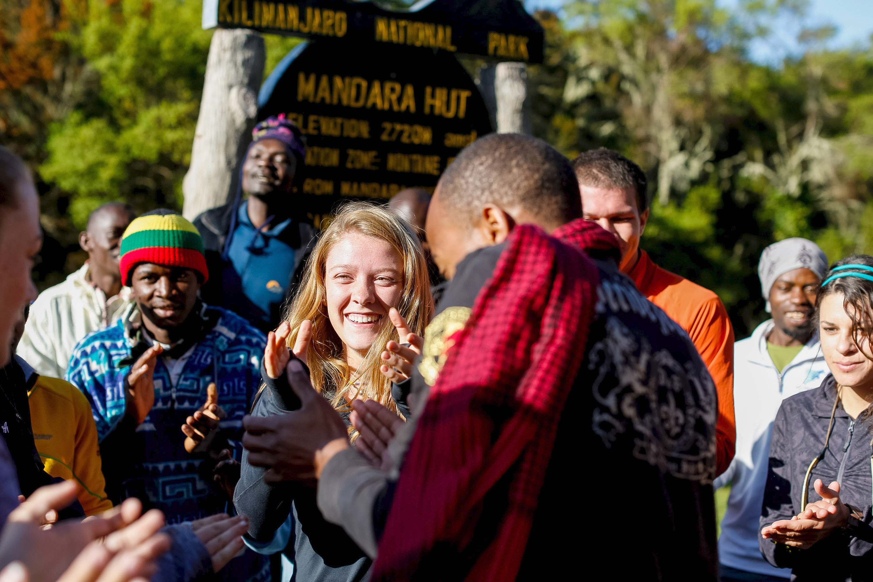 A student smiles amid cheering guides near the Mandara Hut sign at Kilimanjaro National Park