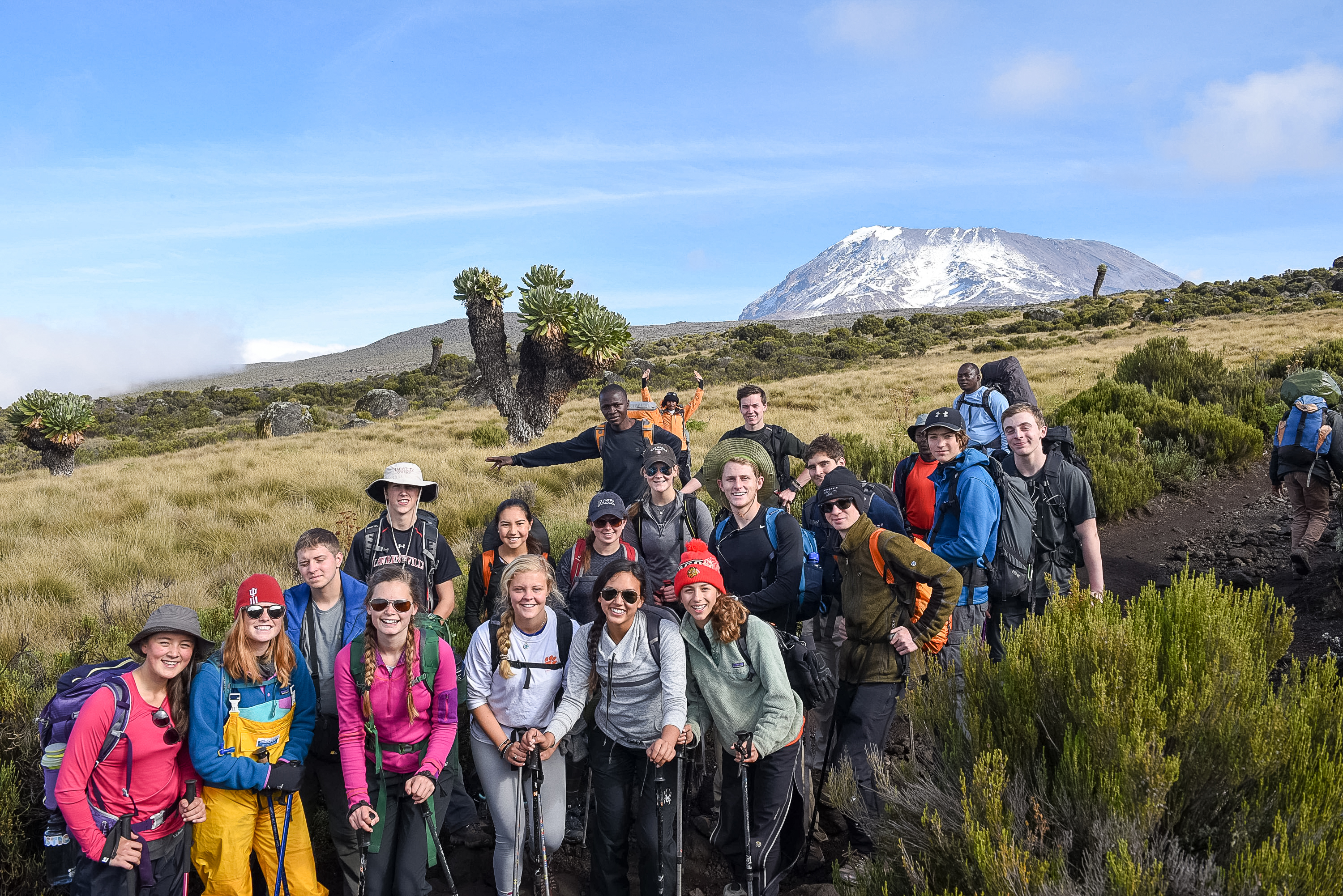 A school group stands together on a grassy plain in Tanzania, with the snow-capped summit of Mount Kilimanjaro rising behind them.