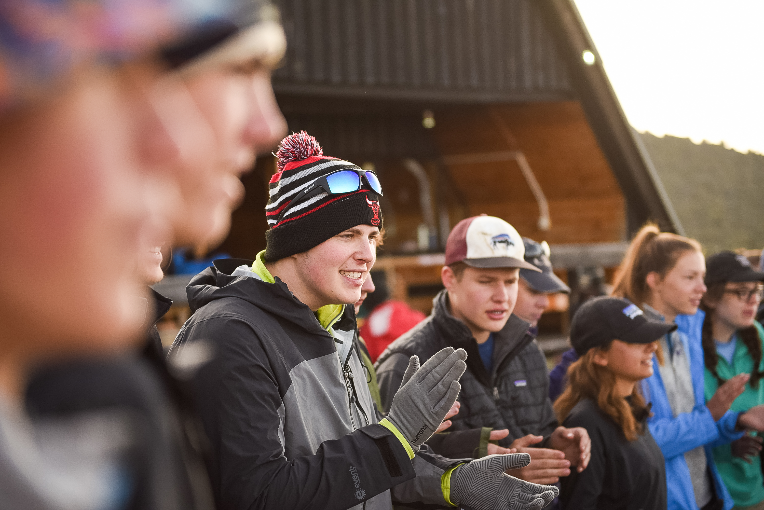 Students in winter hats and jackets clap while standing outdoors near a lodge