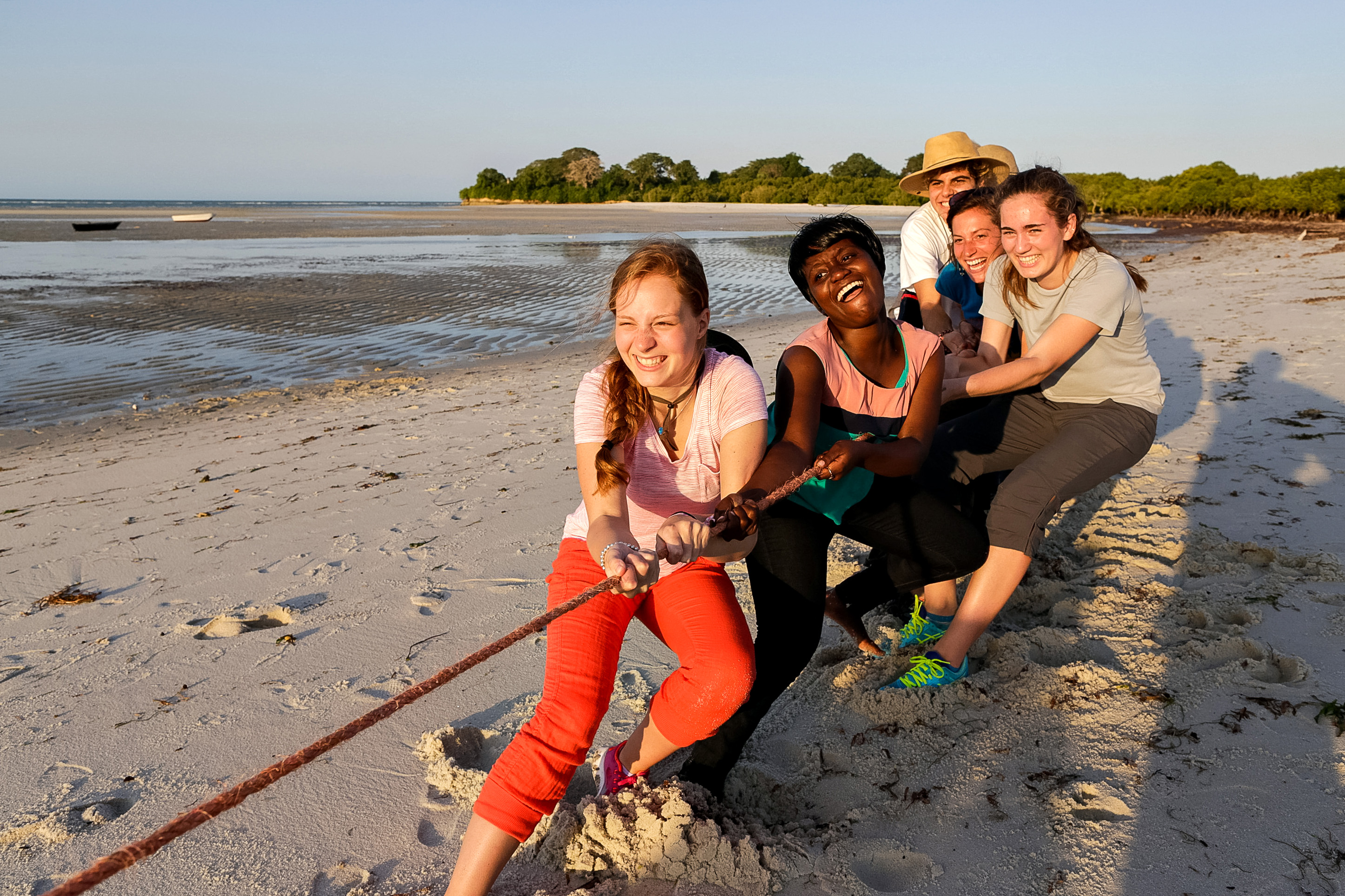 Students pull together on a rope while laughing on a beach at dusk