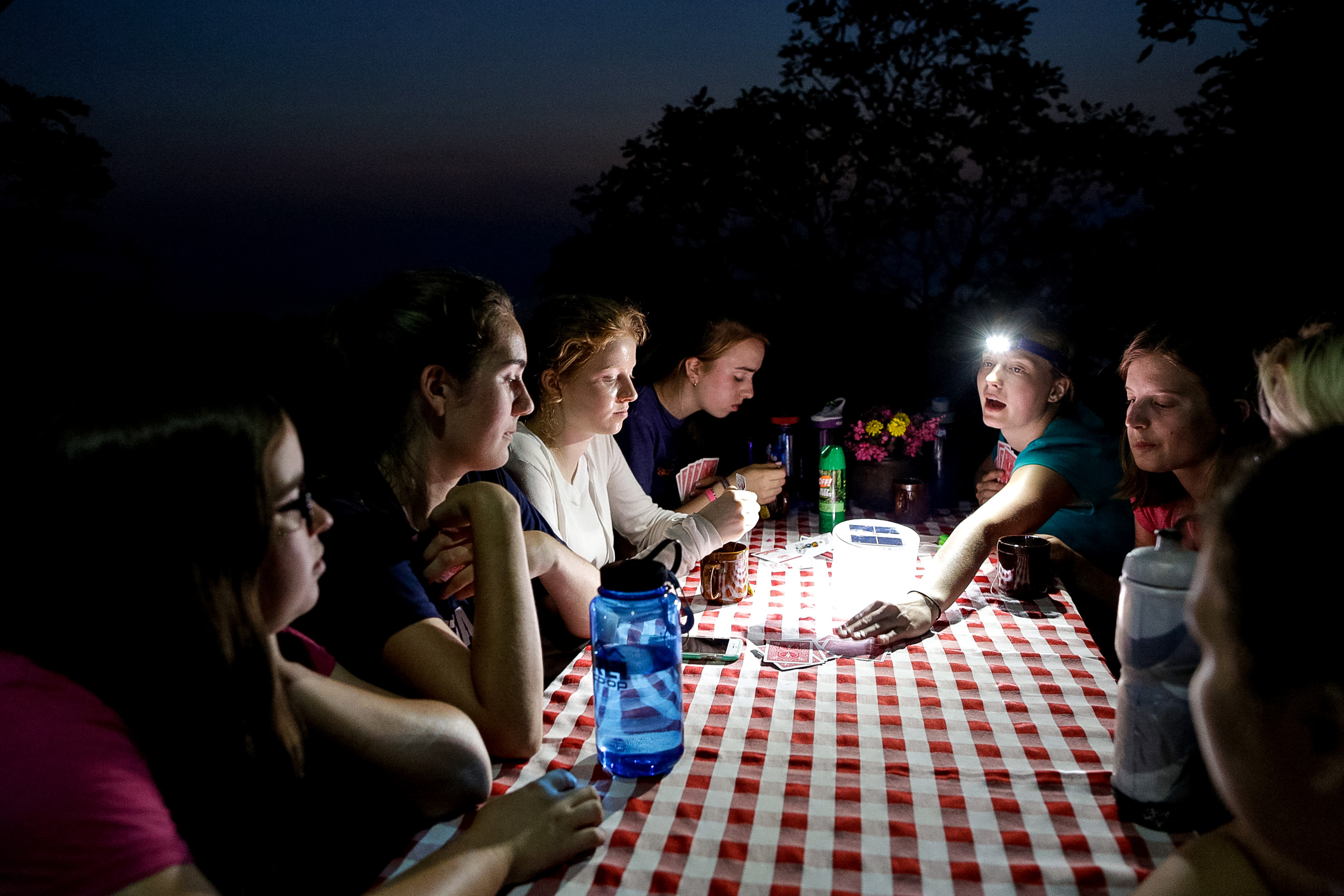 Teens gather around a checkered tablecloth by headlamp light at night
