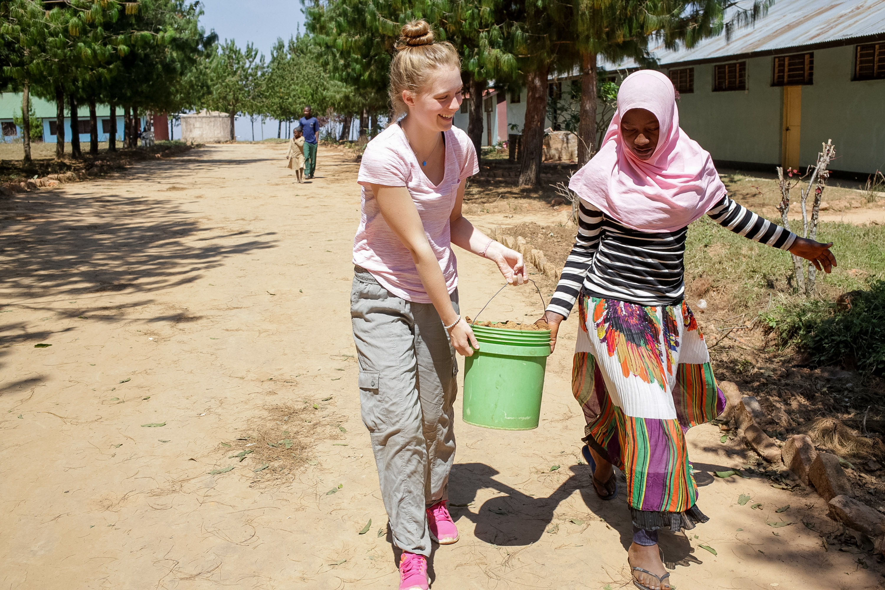 A student and a local girl carry a bucket together along a dirt path