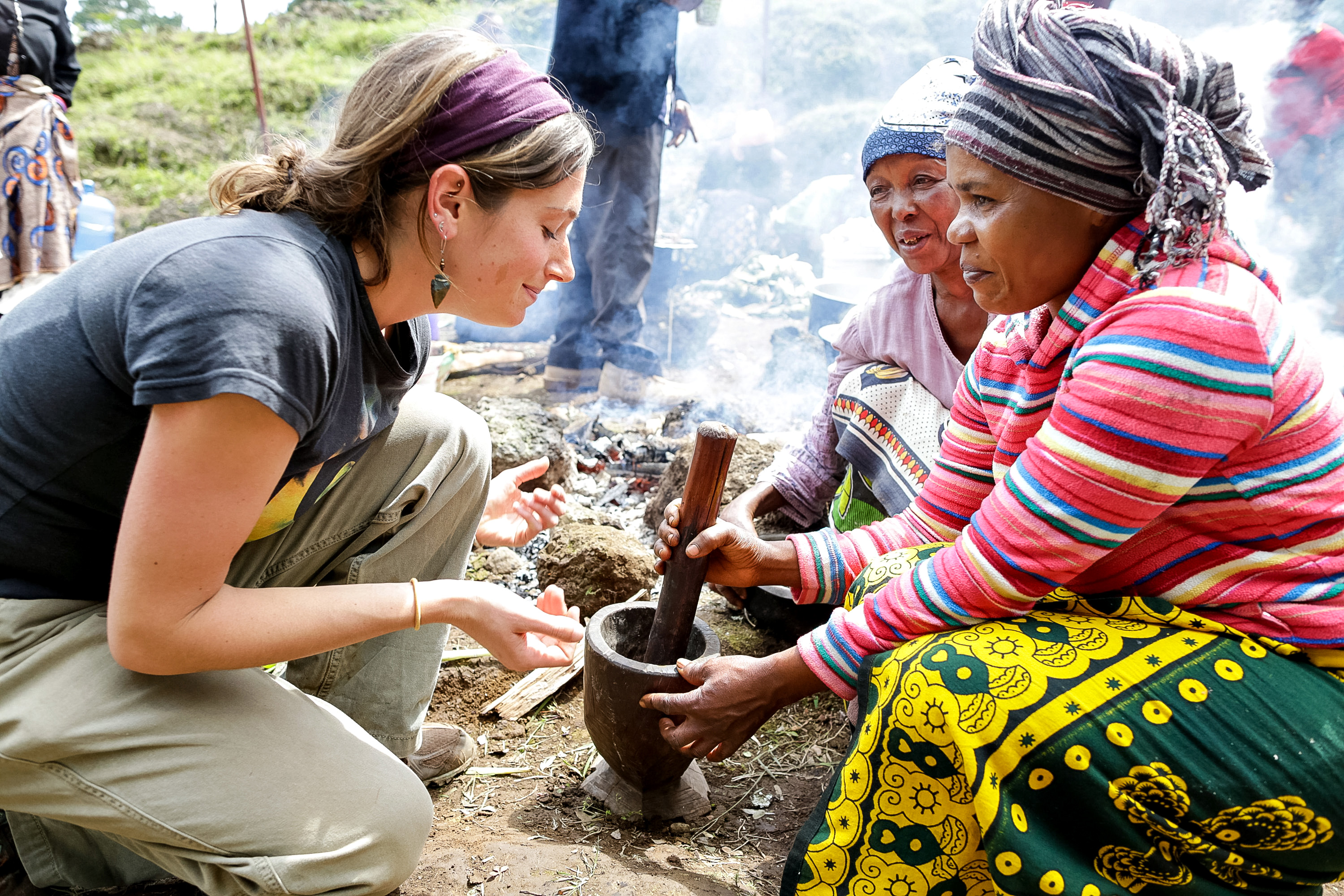 A school trip stops to let a student use a mortar and pestle alongside local women gathered around a fire