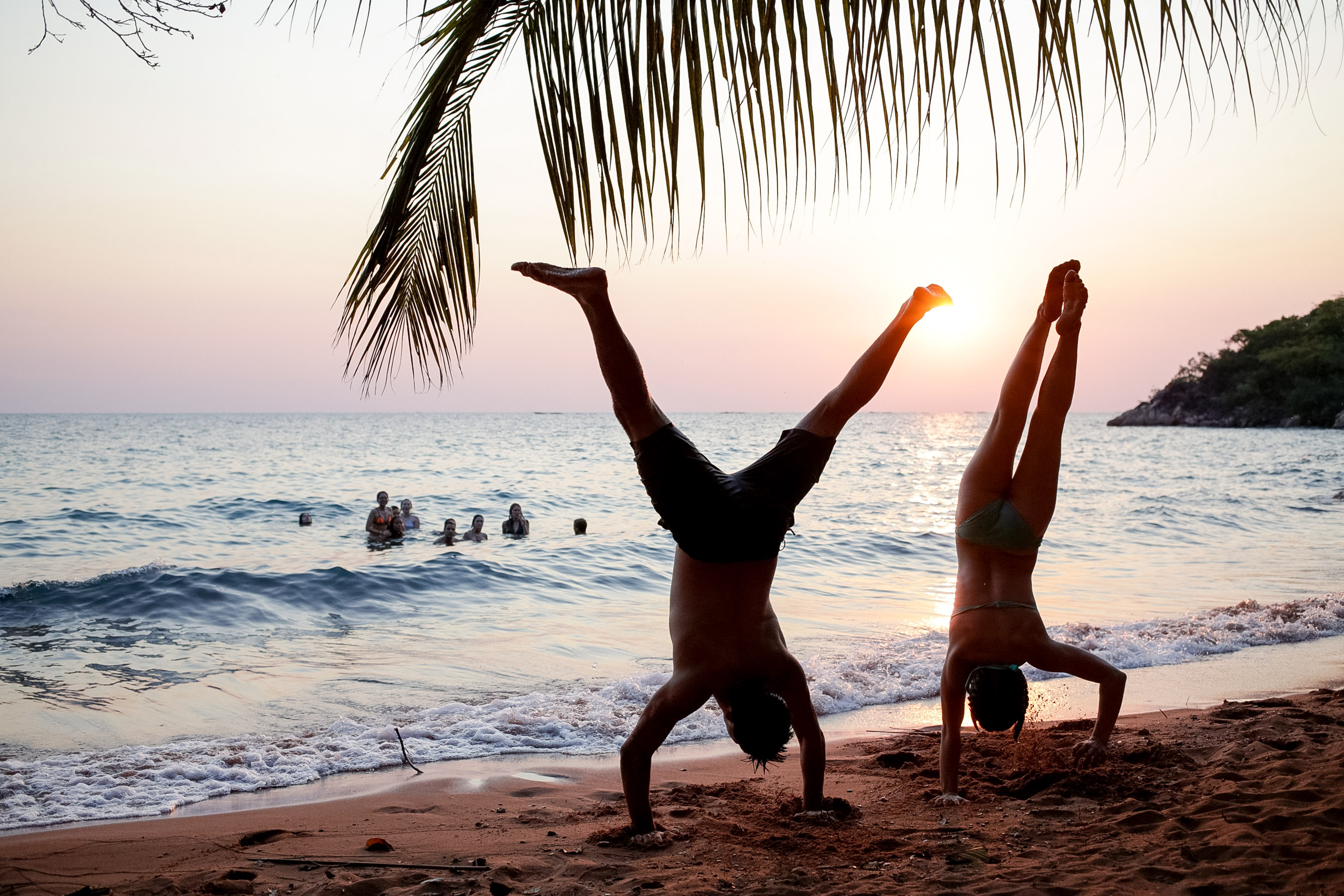 Students do handstands on the beach at sunset while others wade in the ocean behind them