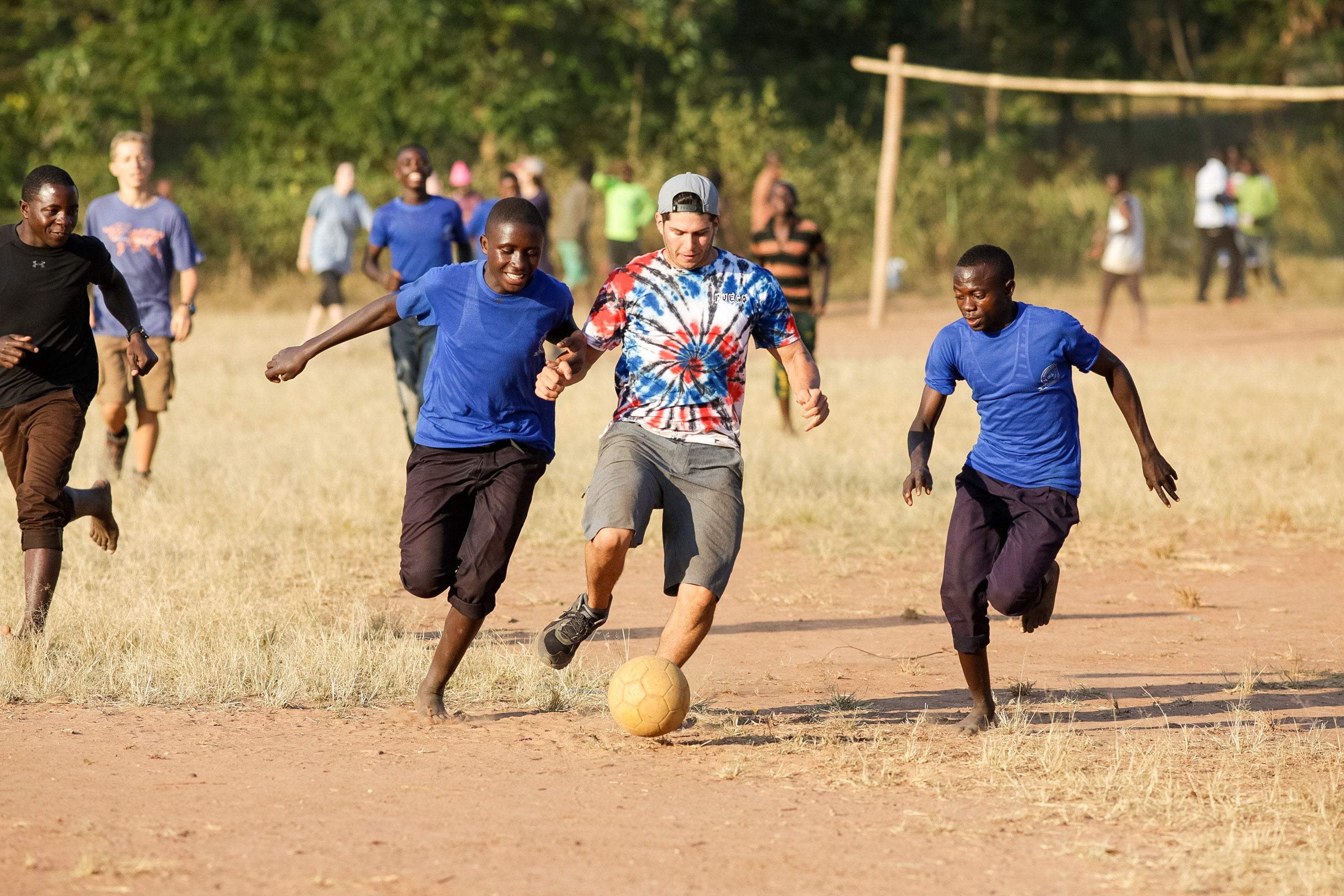 Students chase a soccer ball on a dusty field with local players