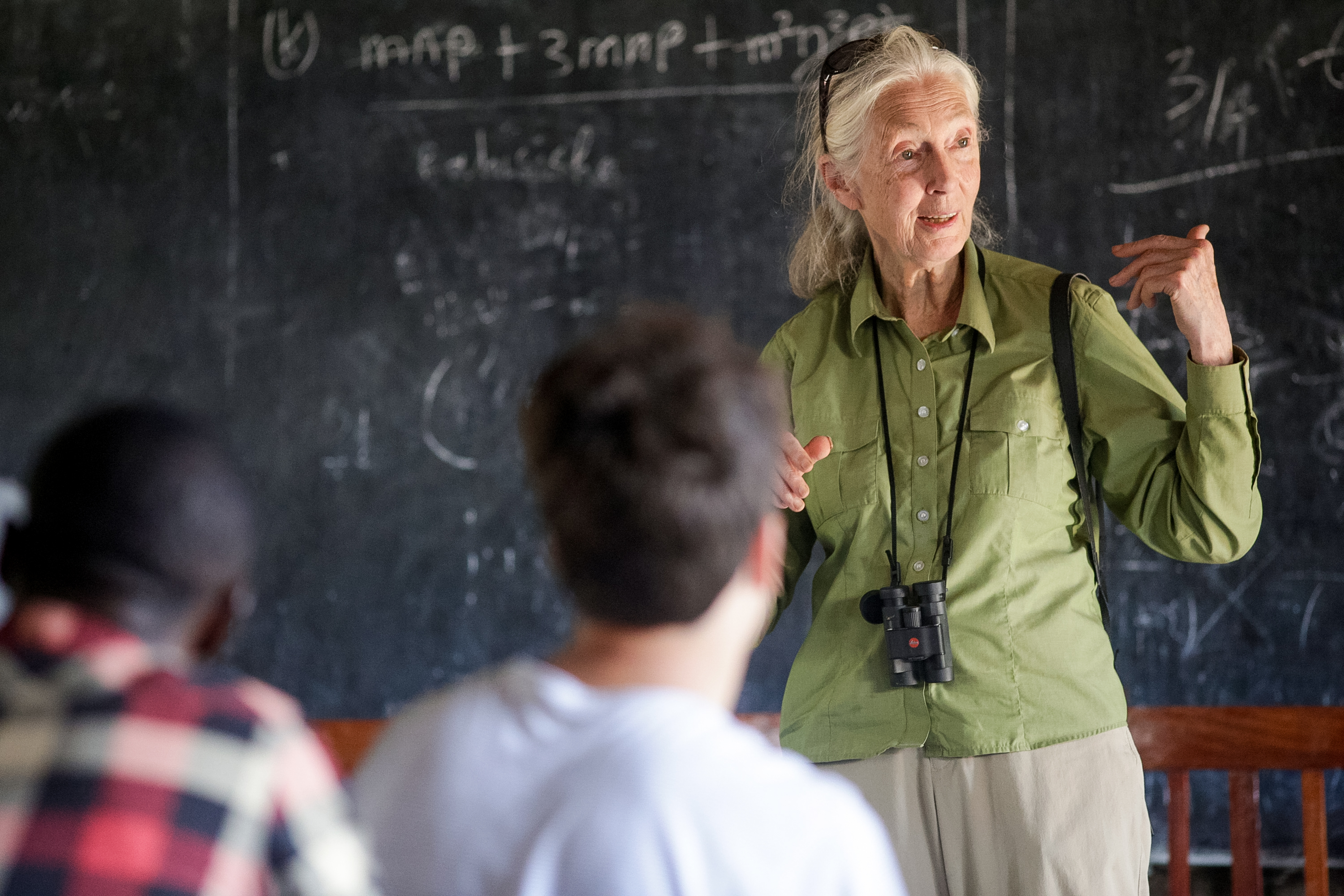 A speaker with binoculars around her neck gestures while teaching students in front of a chalkboard