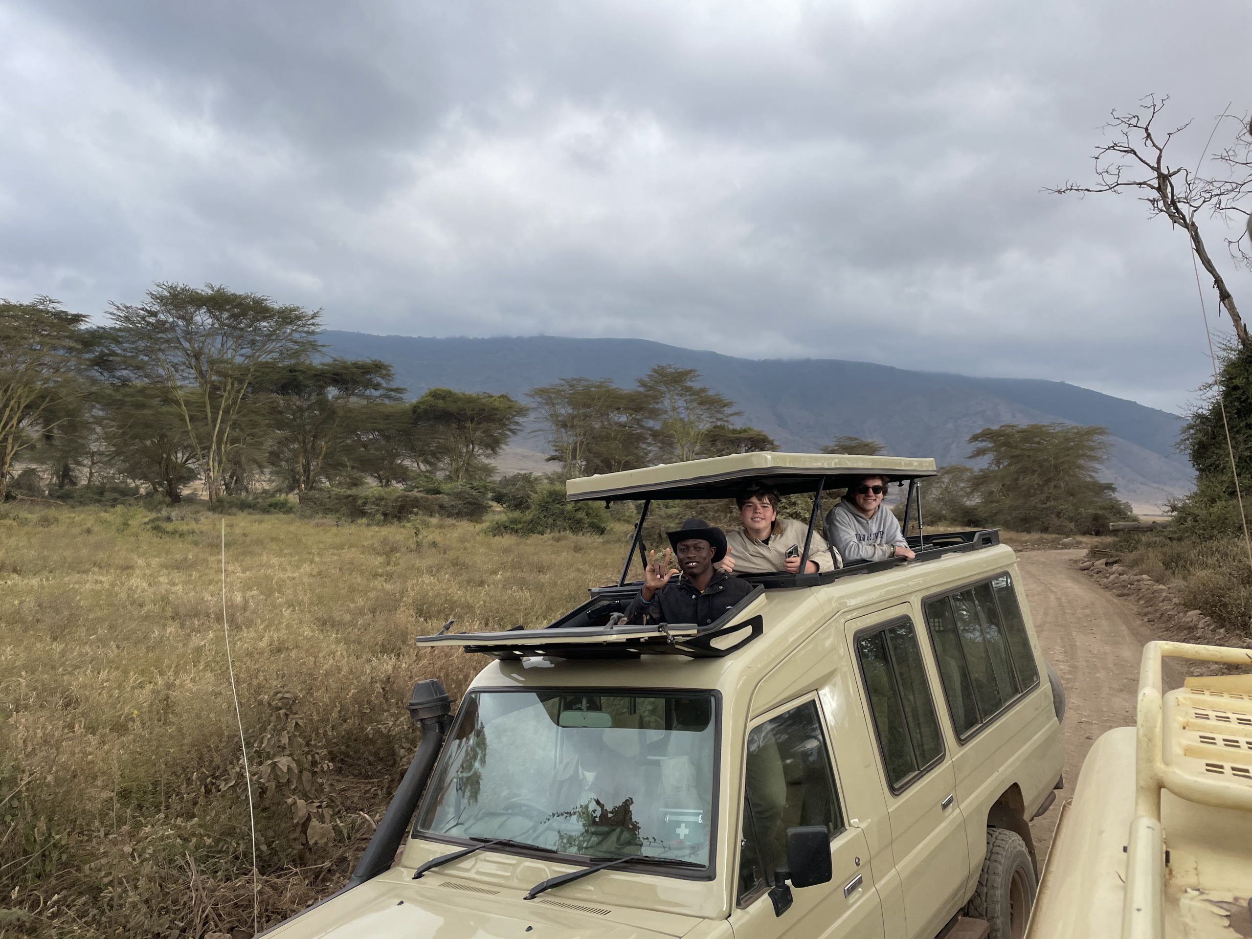 Students look out from the open roof hatch of a safari vehicle parked in dry grassland.
