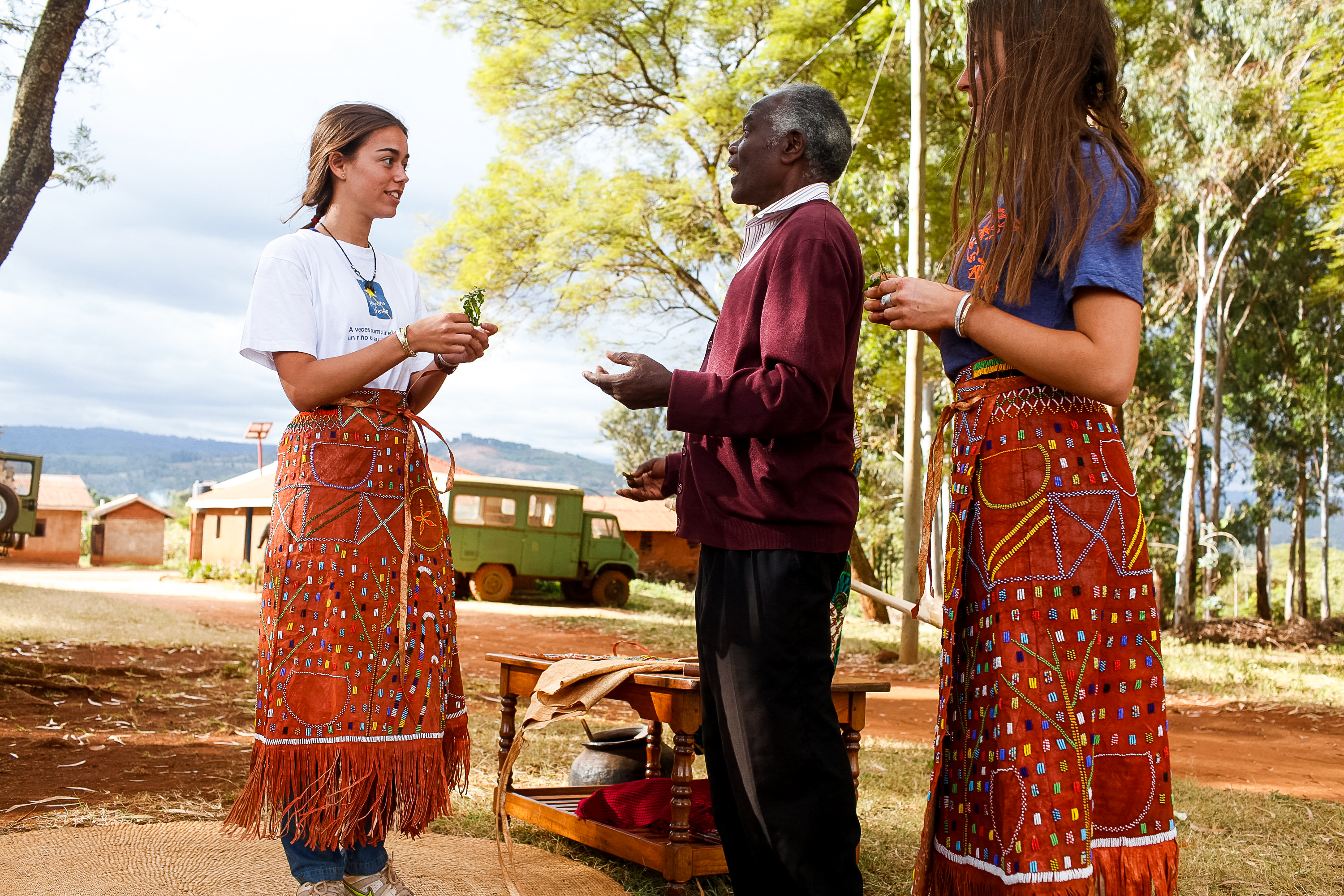 Students wearing traditional beaded skirts talk with a village elder outdoors