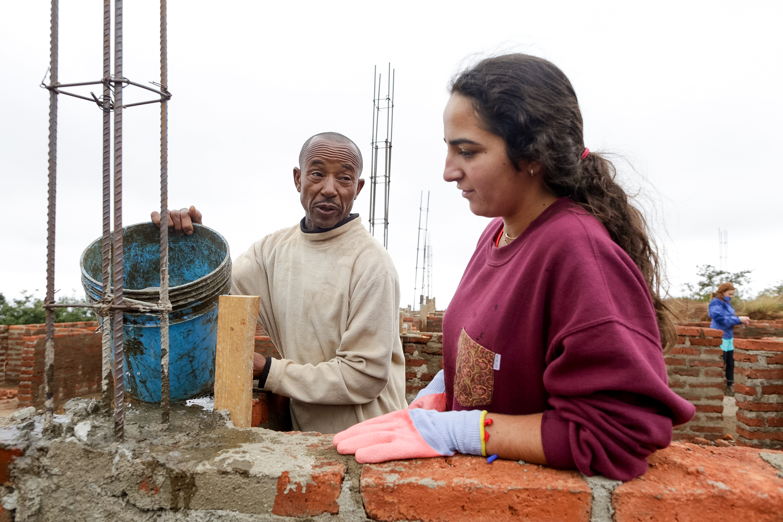 A student in work gloves helps lay bricks alongside a local worker on a construction site