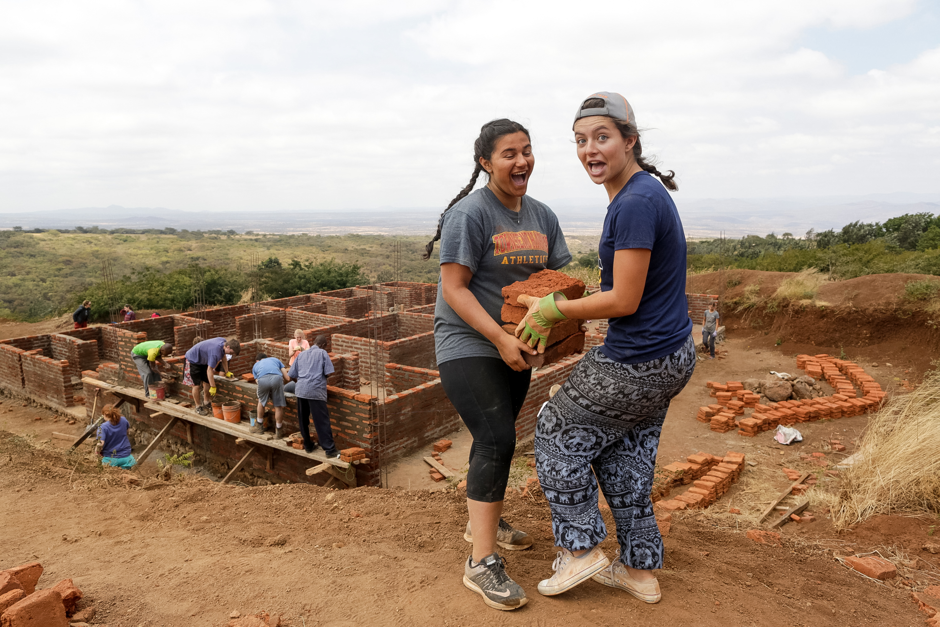 Two students carry a concrete block along a half-built wall on a village construction site, with stacked bricks and timber scaffolding behind them.