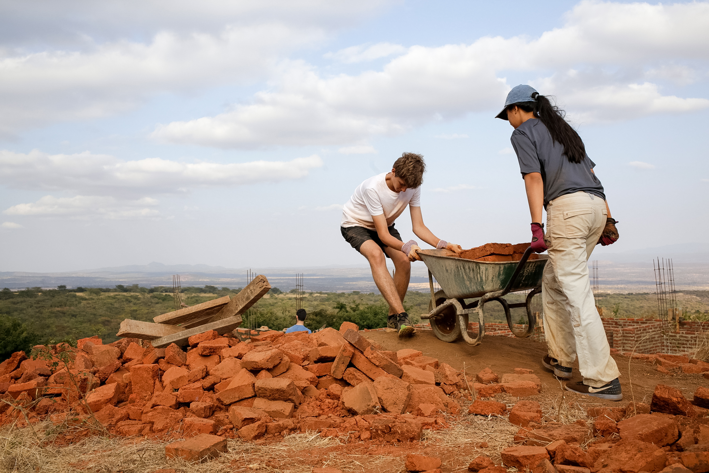 Students load bricks into a wheelbarrow on a hilltop construction site