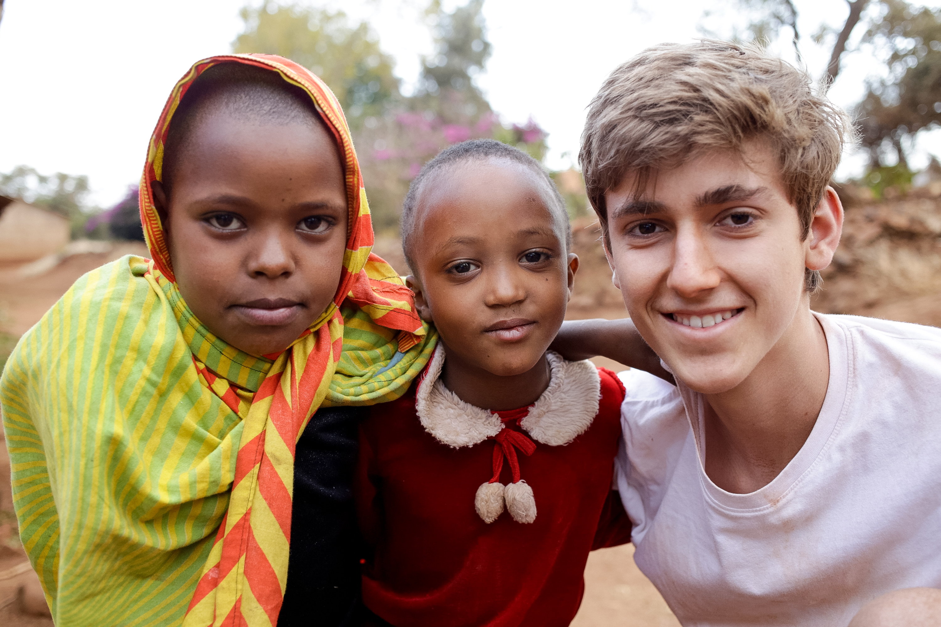 A student stands between two young girls in bright headscarves outside a village school in Tanzania.