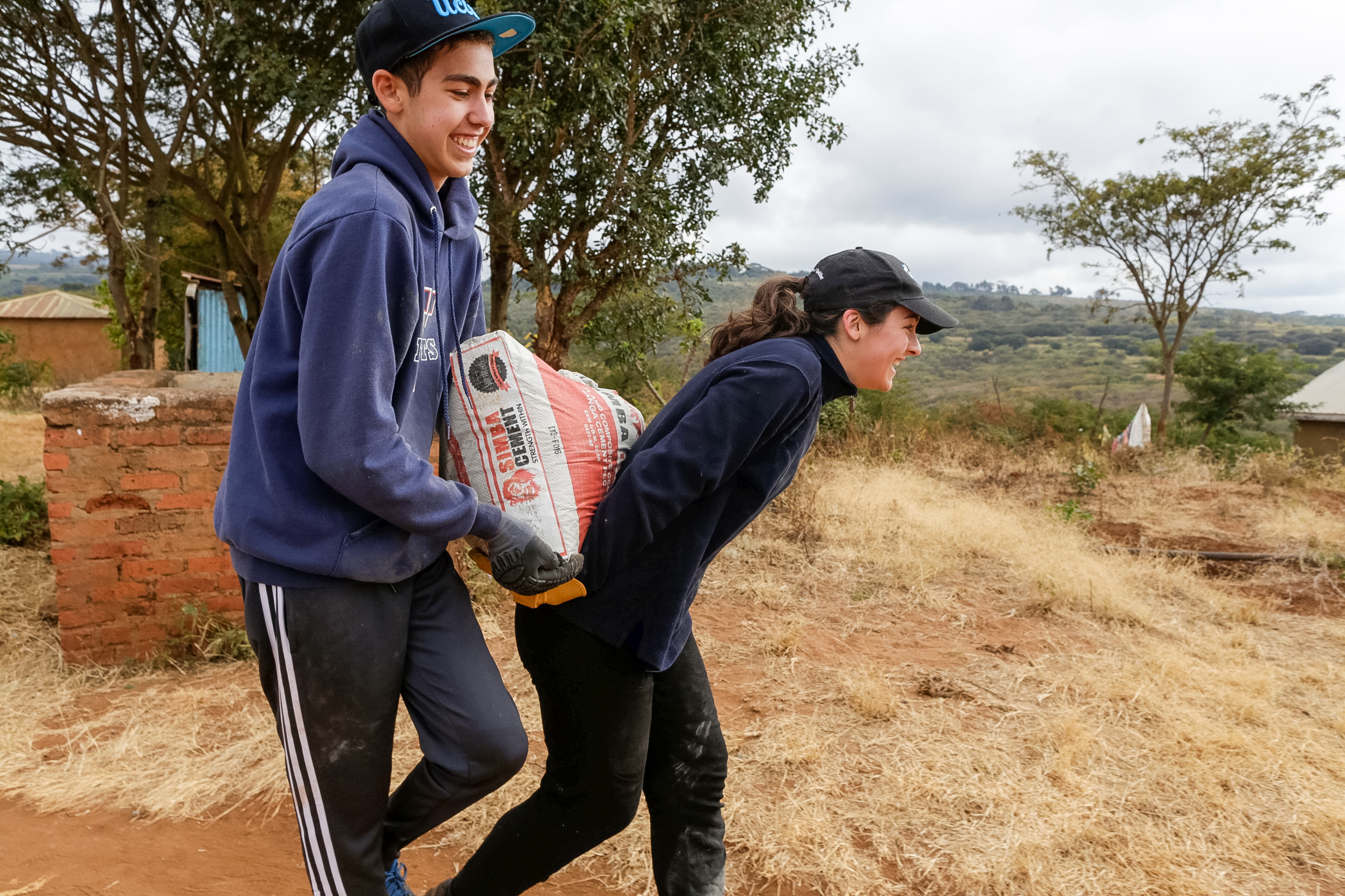 Students carry a bag of supplies together along a dirt path in a village