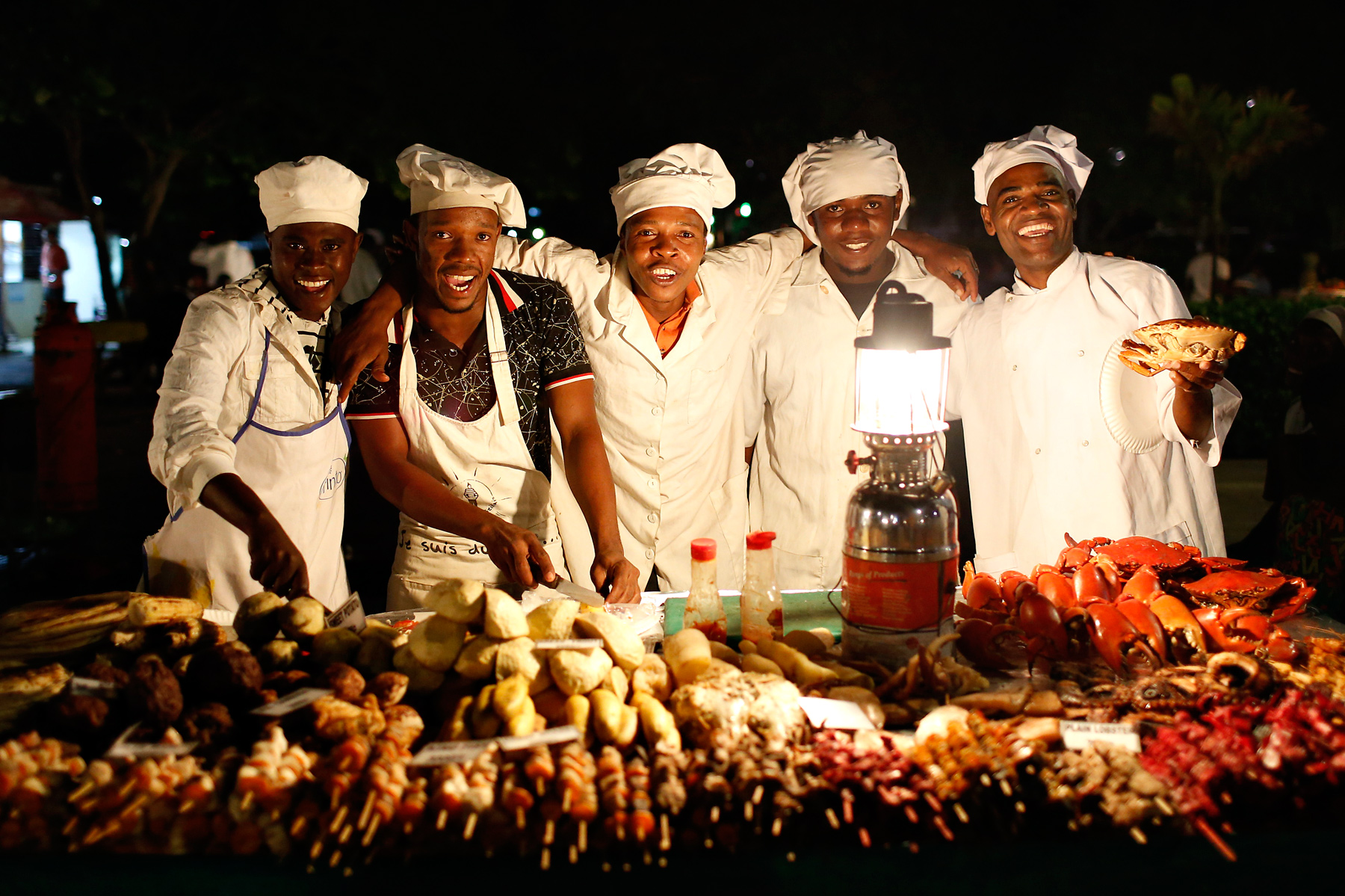 Chefs in white uniforms pose behind a lantern-lit spread of grilled seafood and skewers at night
