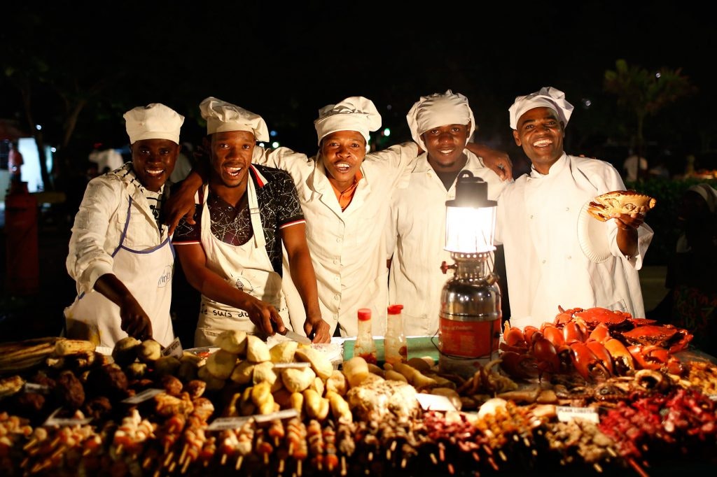 Group of chefs smiling and posing behind a table of food at night.