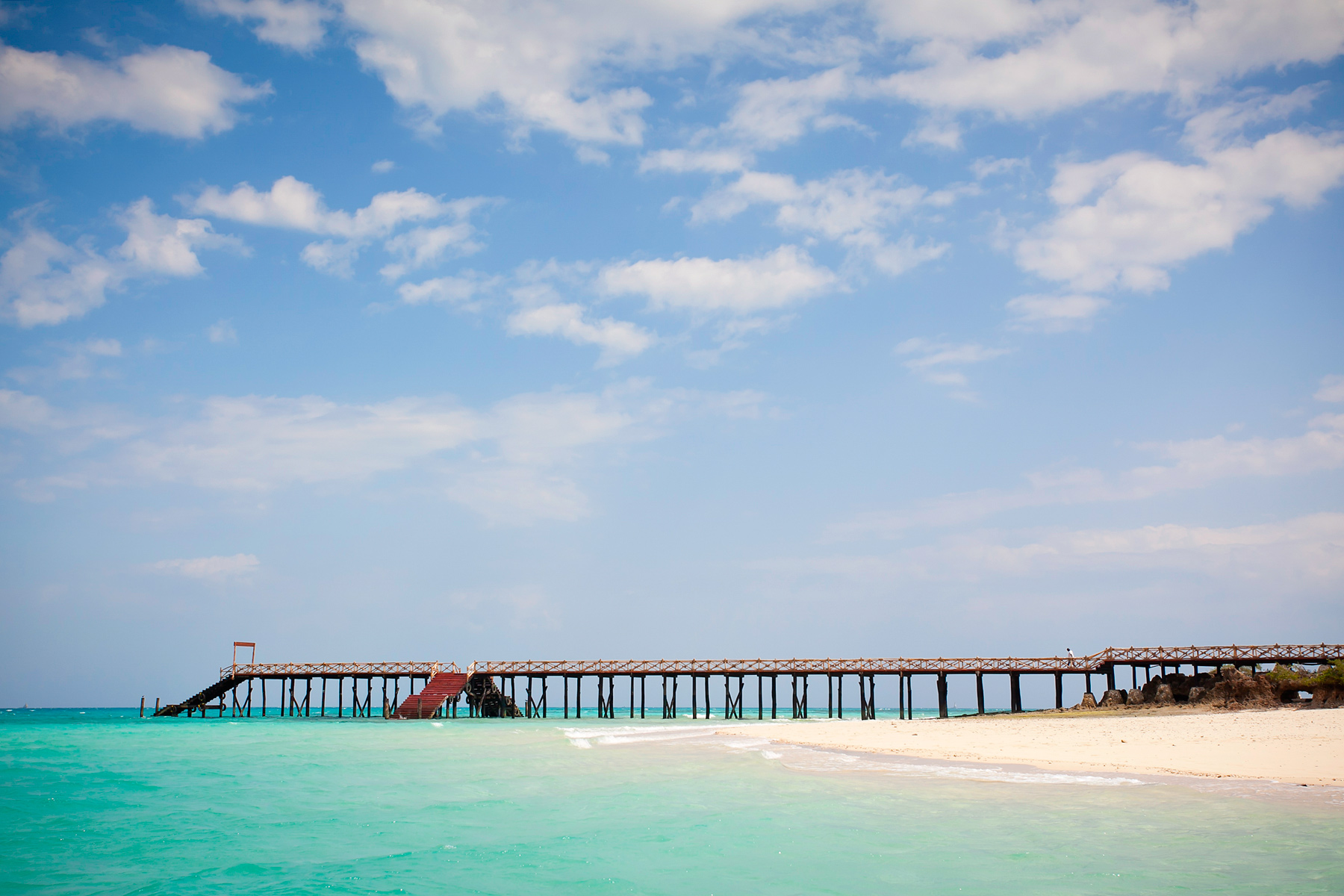 A wooden pier stretches over turquoise water toward a sandy beach