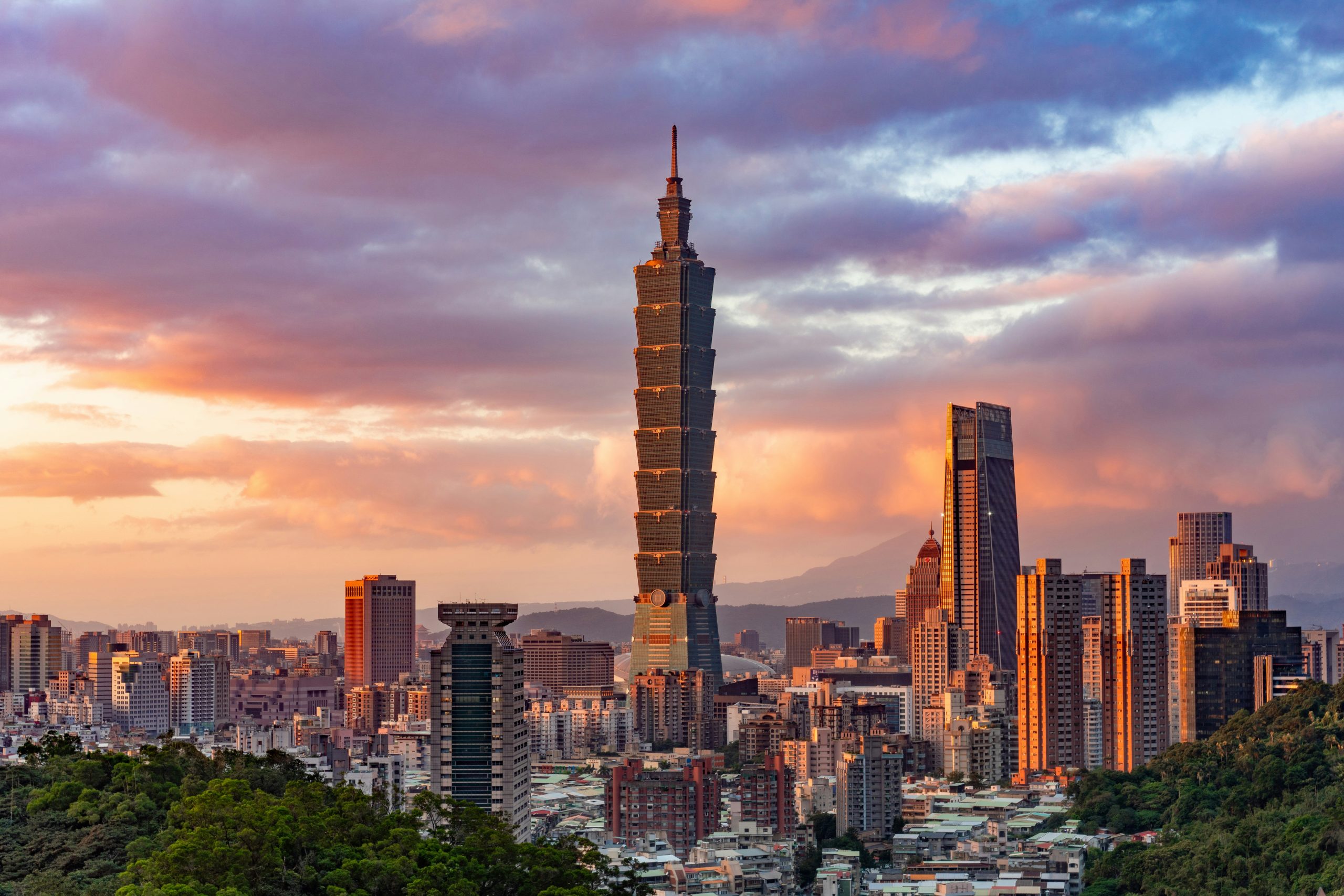 Taipei 101 rising above the Taipei skyline at sunset, with forested hills in the foreground.