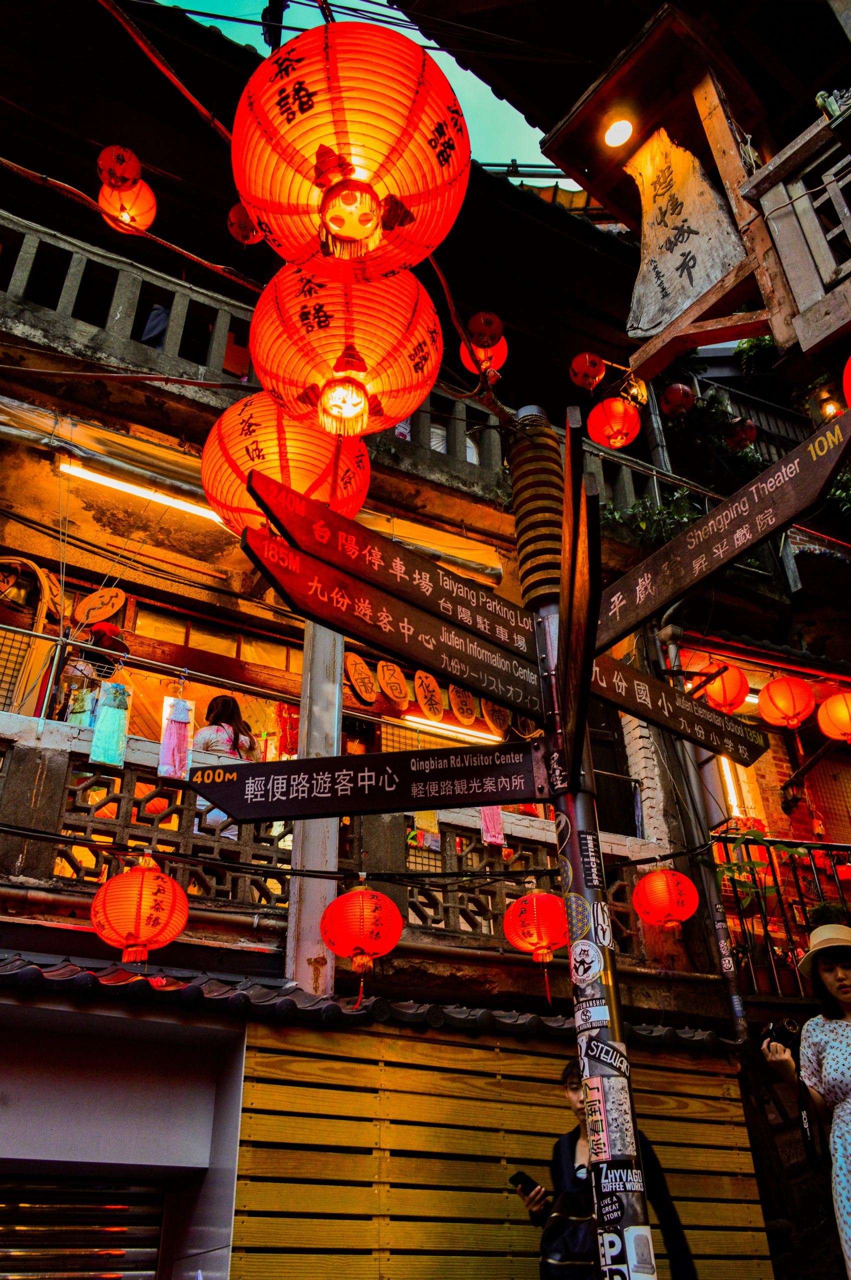 Direction signs and red lanterns at night on a Jiufen street, pointing to the Shengping Theater and the visitor center.