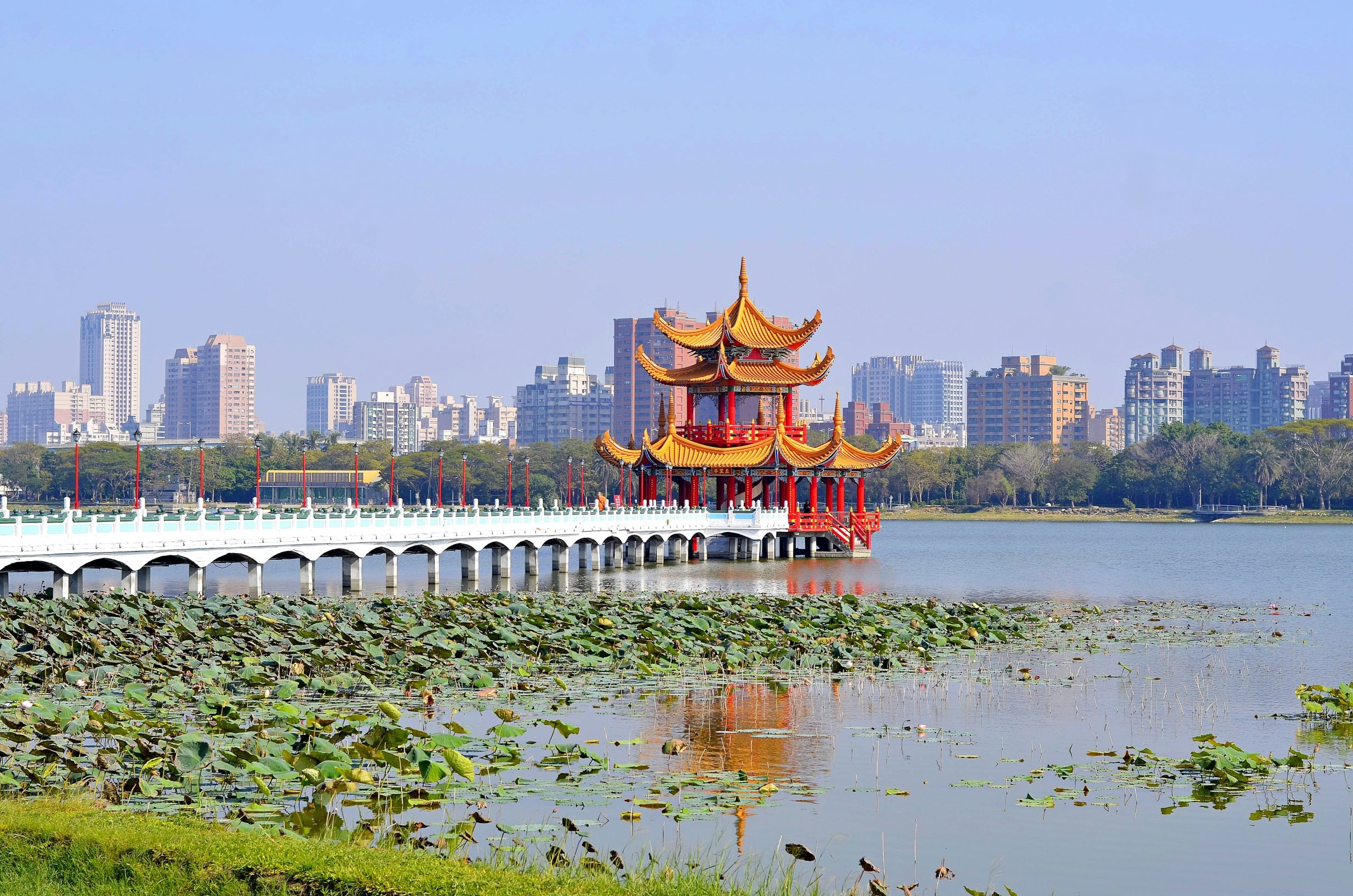 A pavilion on Kaohsiung's Lotus Pond, reached by a long white bridge over lotus leaves, with the city skyline behind.