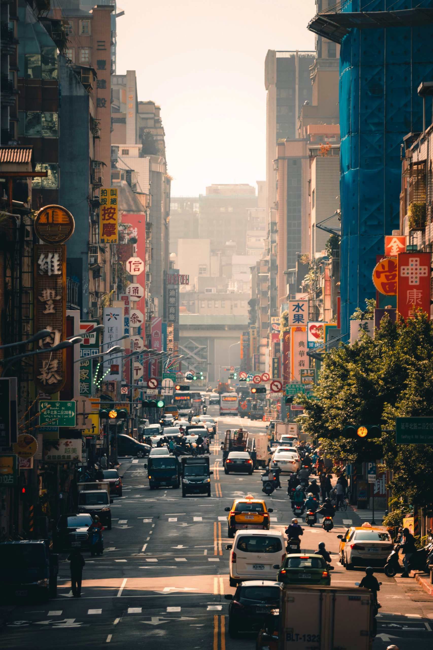 A busy Taiwanese city street lined with vertical shop signs, with taxis, cars, and scooters heading toward high-rise buildings.