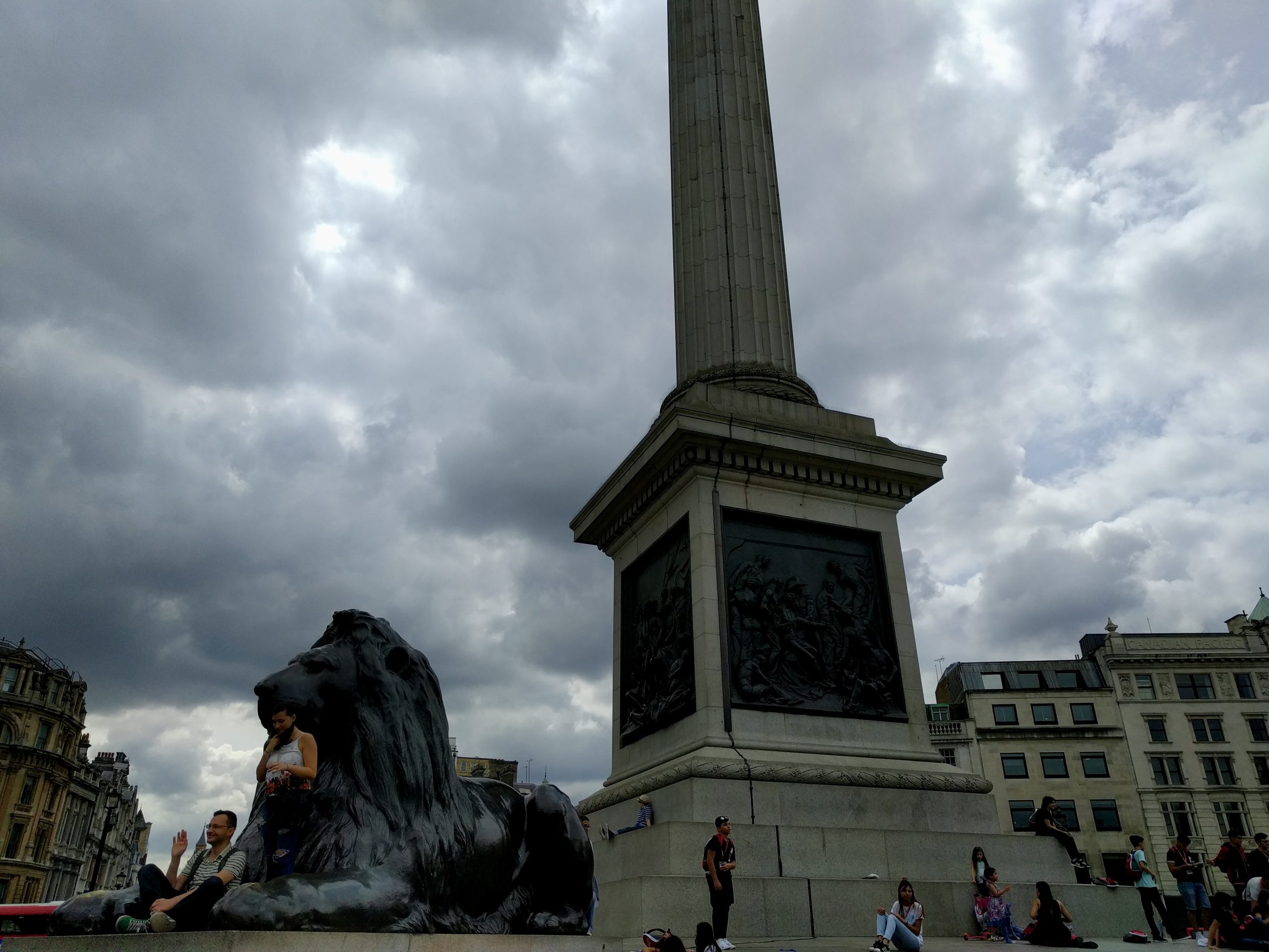 Nelson's Column rises beneath a cloudy sky as visitors sit near the bronze lion statue at its base