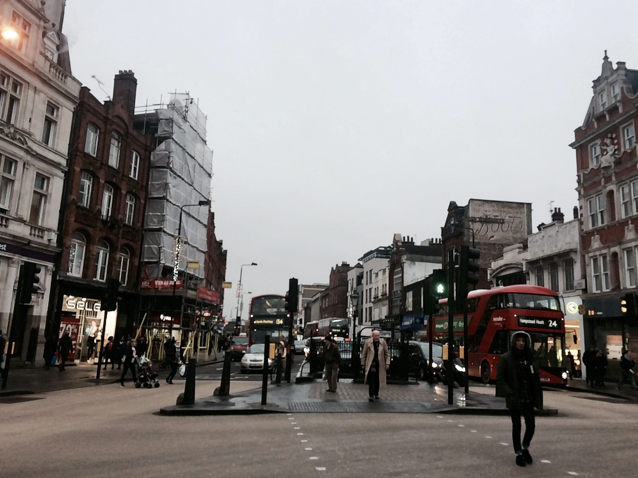 A London street intersection with a red double-decker bus and pedestrians crossing under an overcast sky