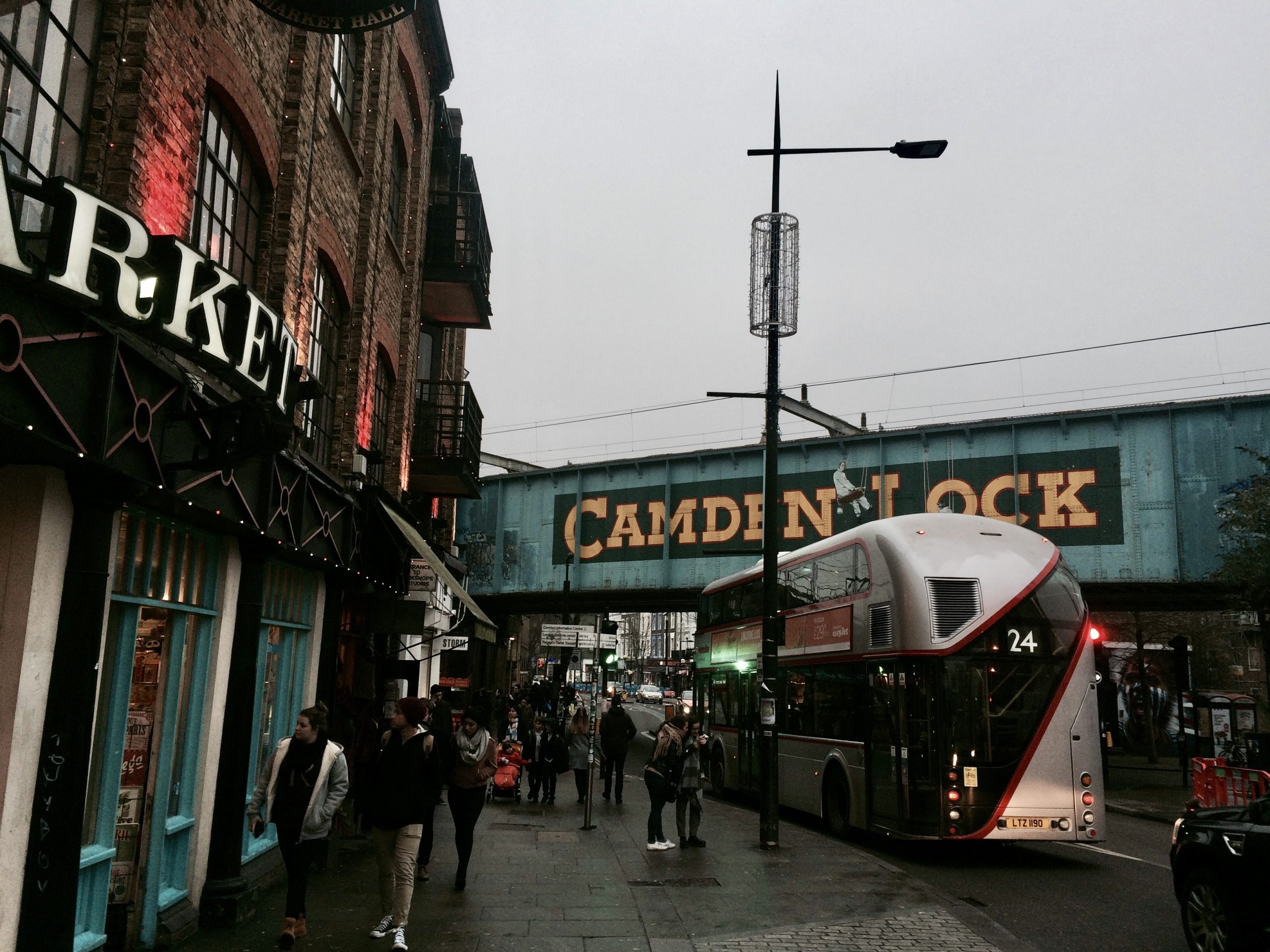 Student travelers watch a double-decker bus pass beneath the Camden Lock railway bridge on a busy London street
