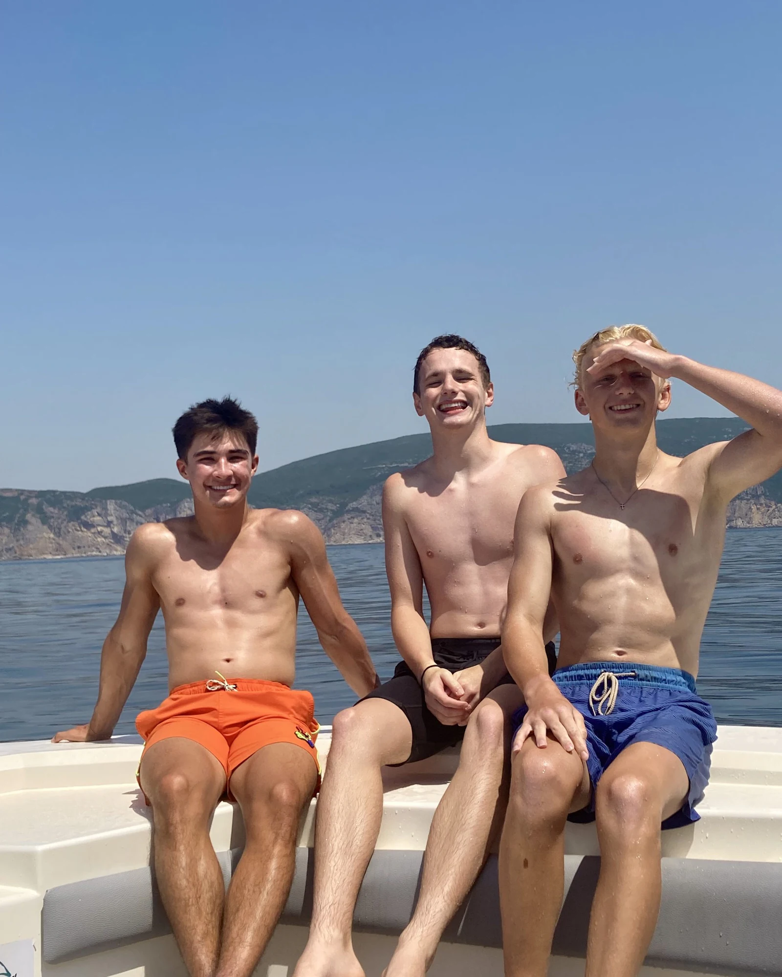 Three teen travelers sit on a boat deck with coastal cliffs and blue sky behind them