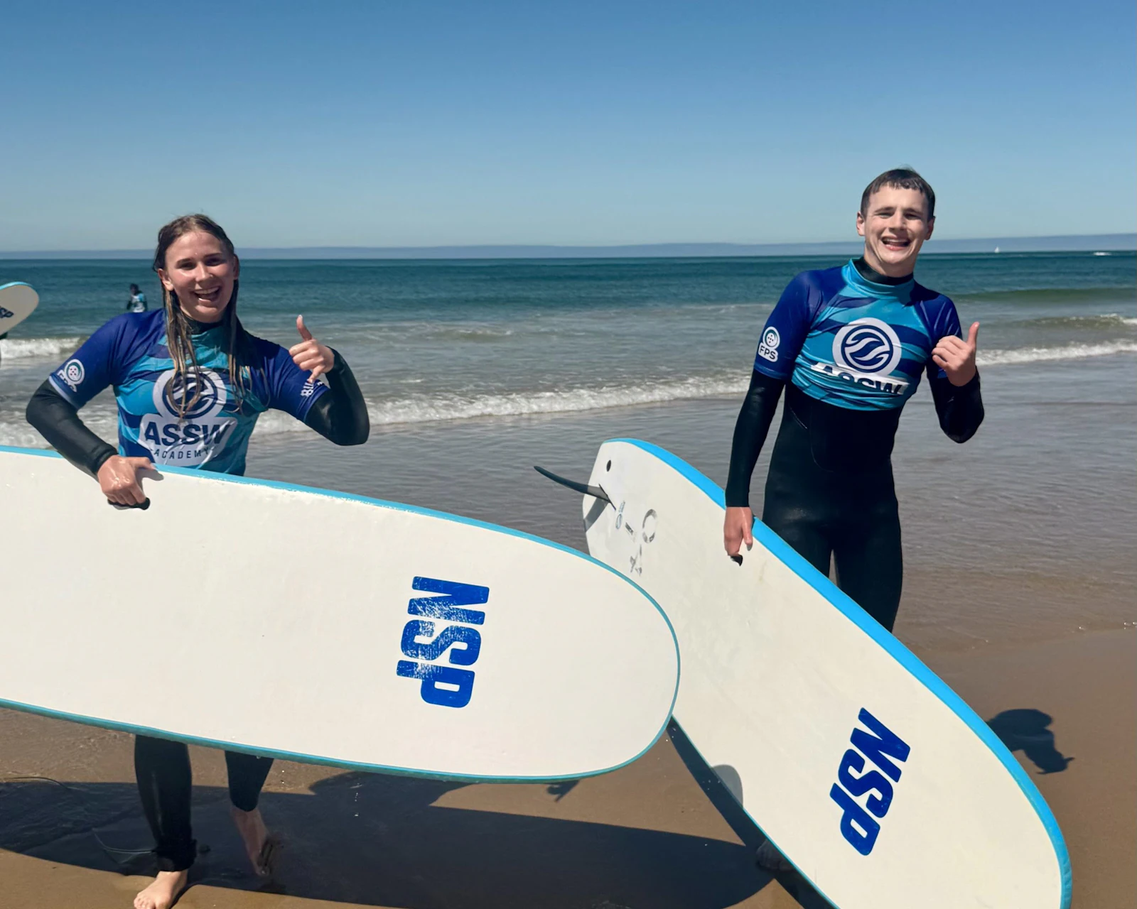 Two teen travelers in rash vests hold surfboards and give a thumbs up on a beach in Portugal