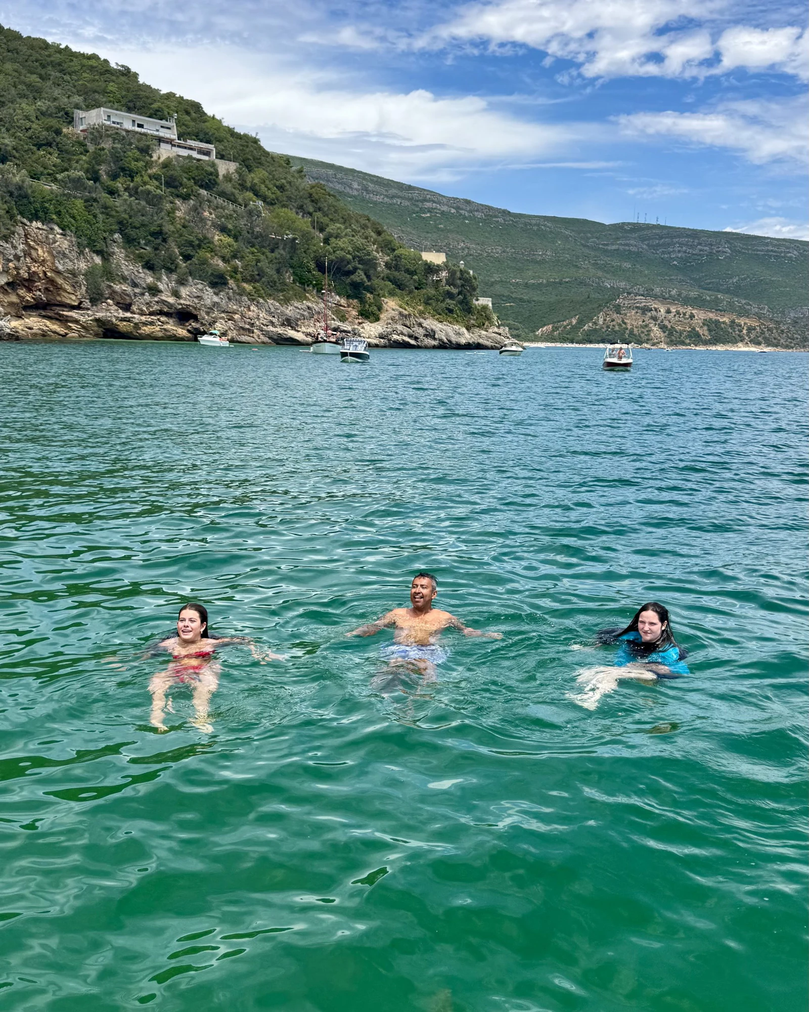 Three teen travelers swim in a sheltered green bay below wooded hills in Portugal, a boat anchored behind them