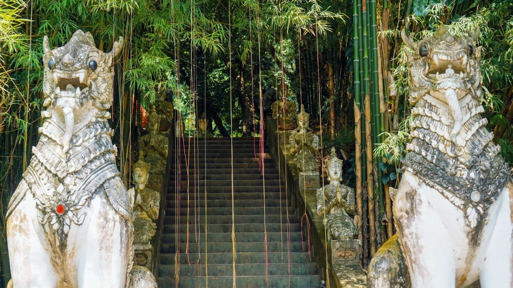 Two statues that look like mythical dogs are at the bottom of a staircase leading up into a temple in the mountains in Thailand.
