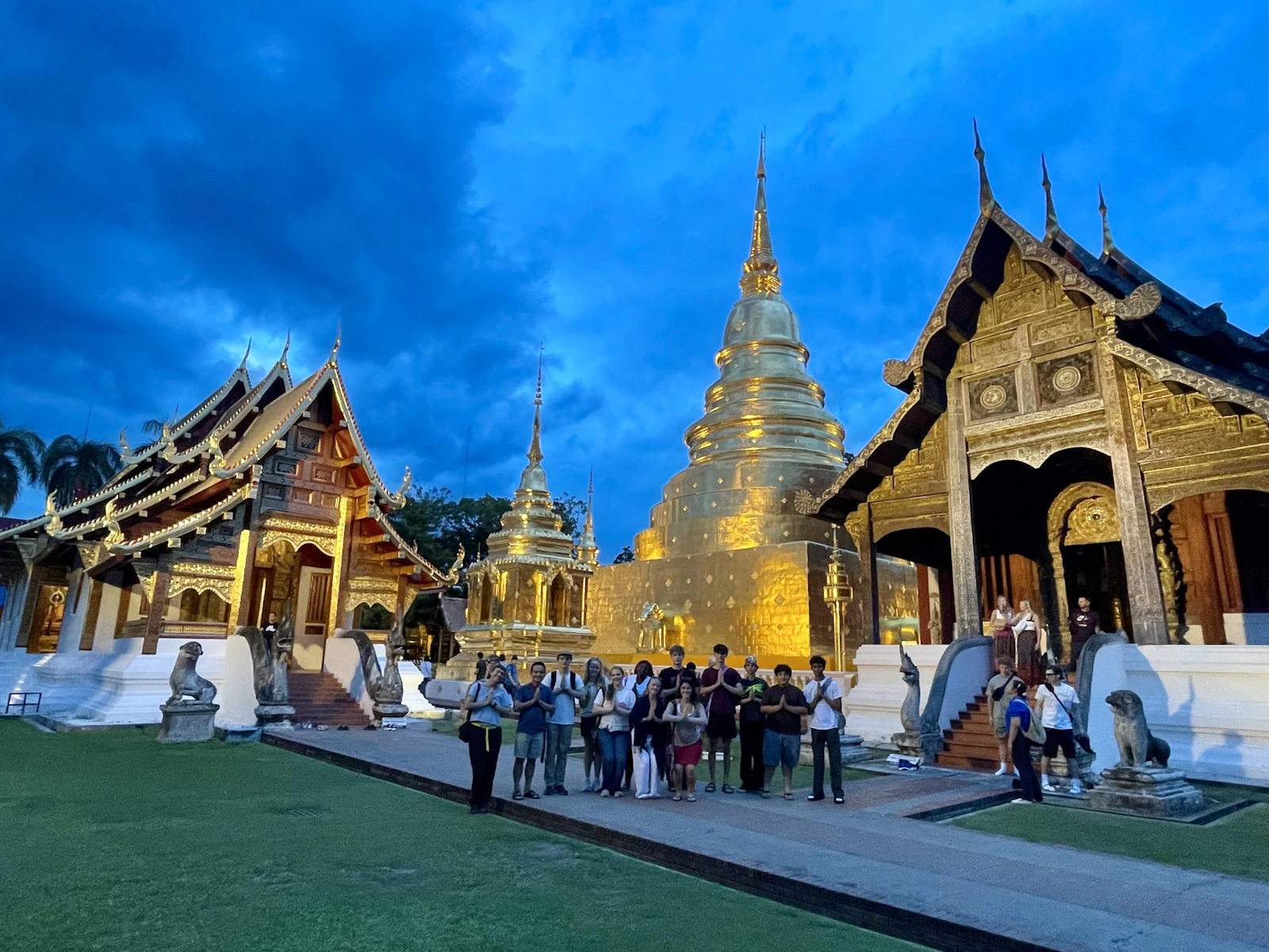 Teen travelers stand in front of a floodlit golden chedi and gilded Lanna temple buildings under a deep blue evening sky in Chiang Mai.