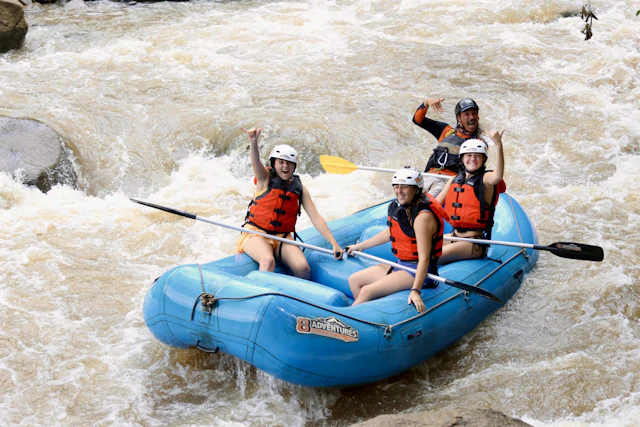 Four teen travelers in helmets and life jackets cheer with paddles raised in a blue raft on brown whitewater in Thailand.