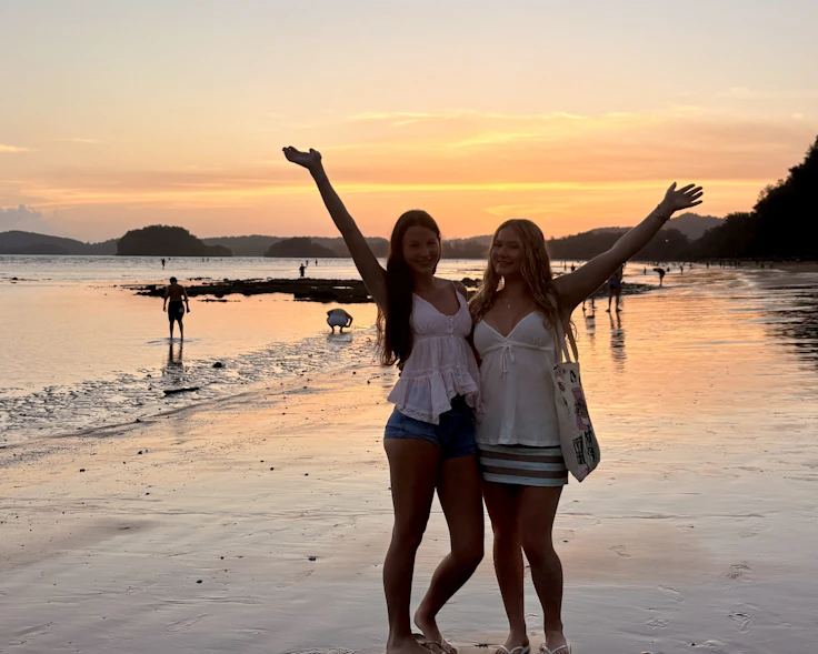 Two teen travelers throw their arms up on a wet Thai beach at sunset, the sky and sand gold behind them.