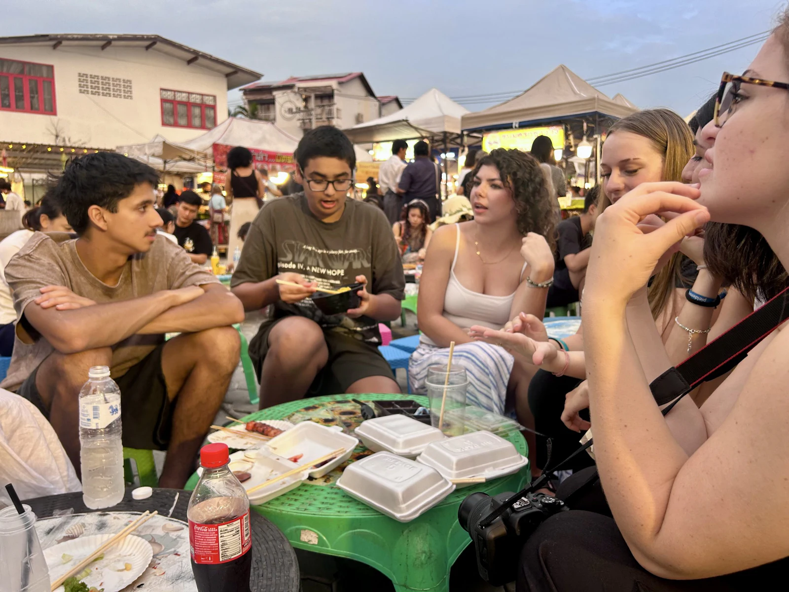 Teen travelers sit around a low green plastic table eating street food at a busy Thai night market at dusk.