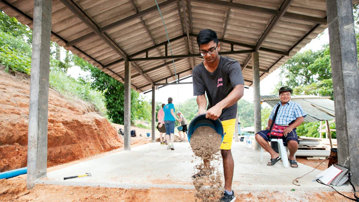 A teen traveler tips a bucket of gravel onto a concrete slab as an older man looks on under an open-sided building shelter.