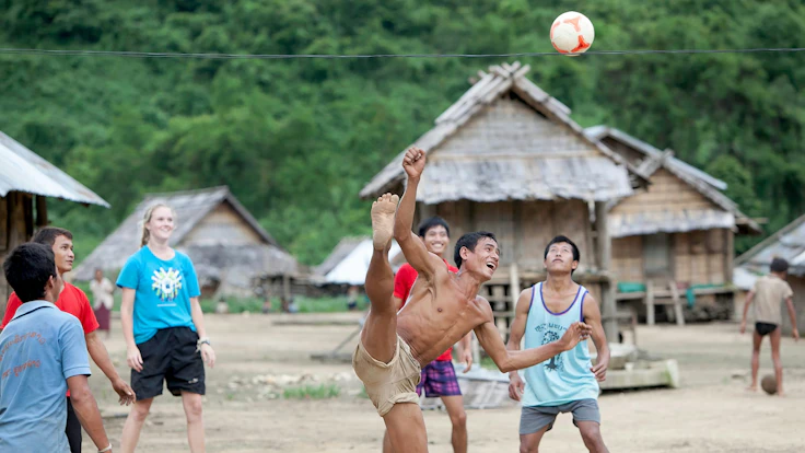 A villager kicks a ball high overhead in a game on a dirt village square while teen travelers and locals watch beside thatched huts.
