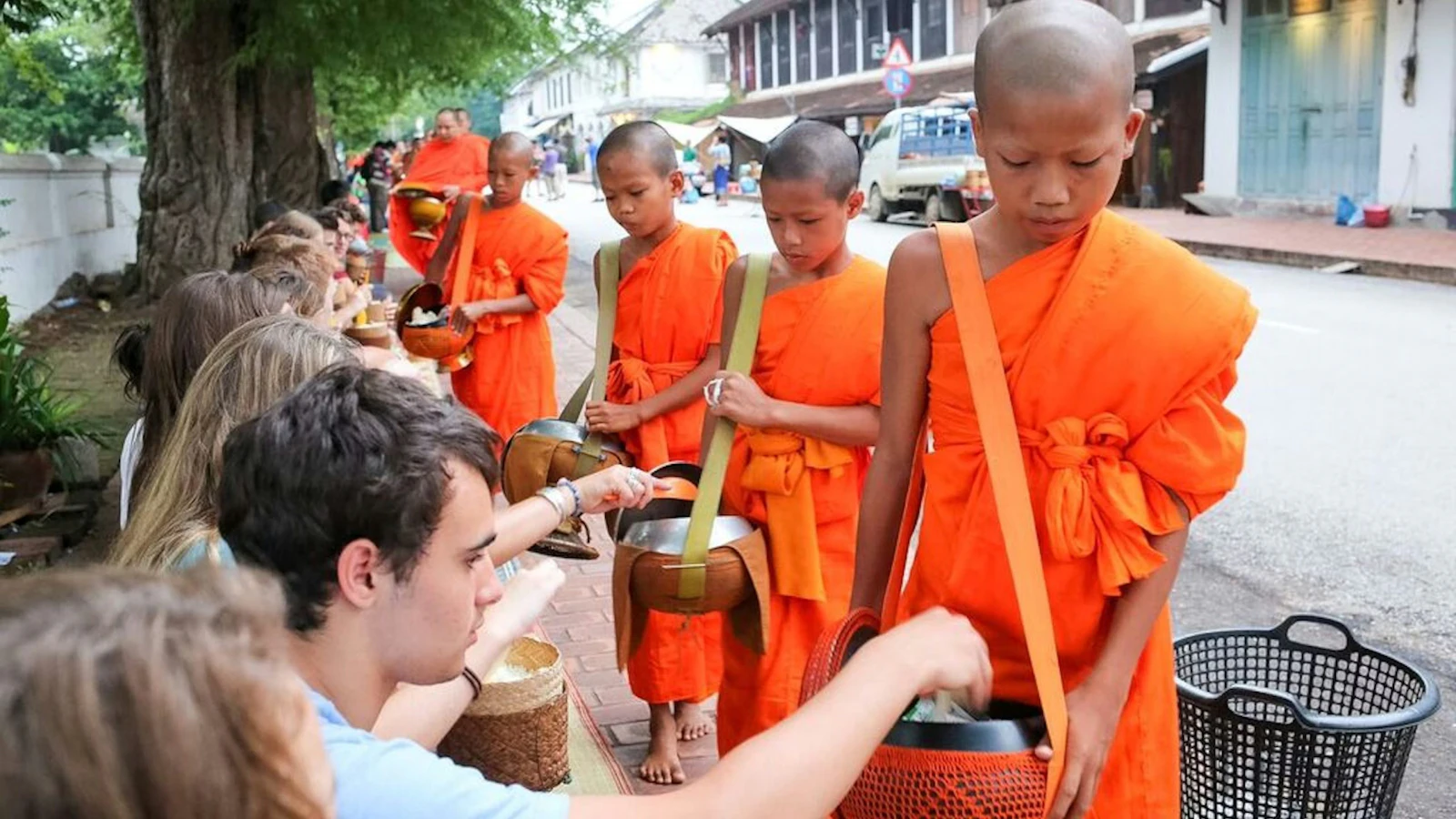 Teen travelers hand food to young monks in orange robes collecting alms on a tree-lined street.