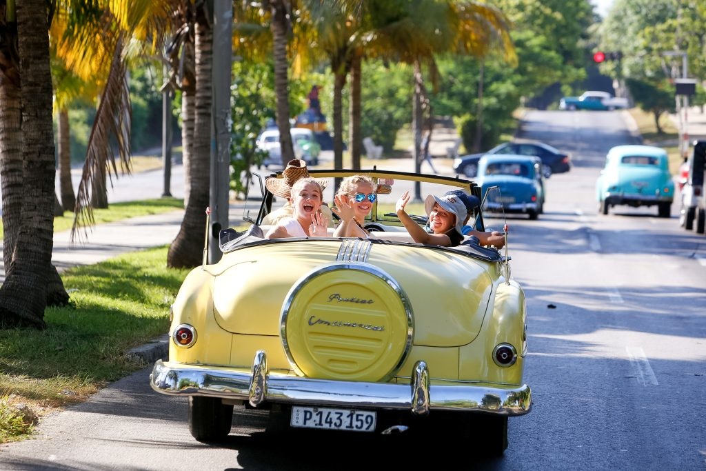 Three teens waving from the backseat of a yellow convertible during an outdoor program in Cuba.